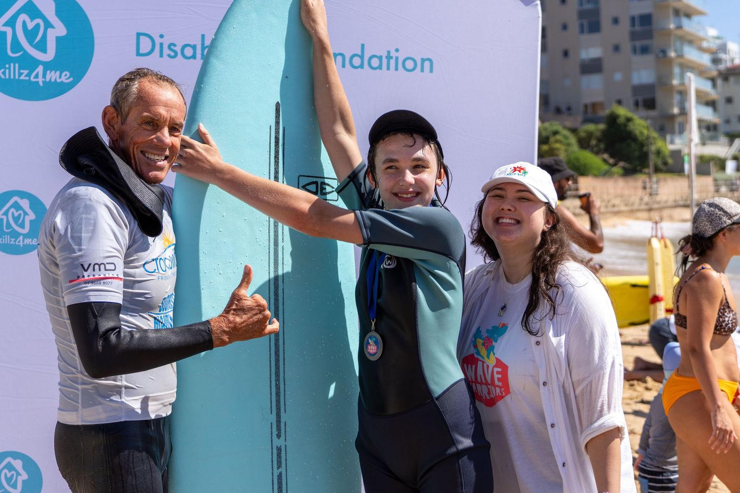 A boy poses with two adults next to a surfboard at a Disability Foundation beach surfing event.