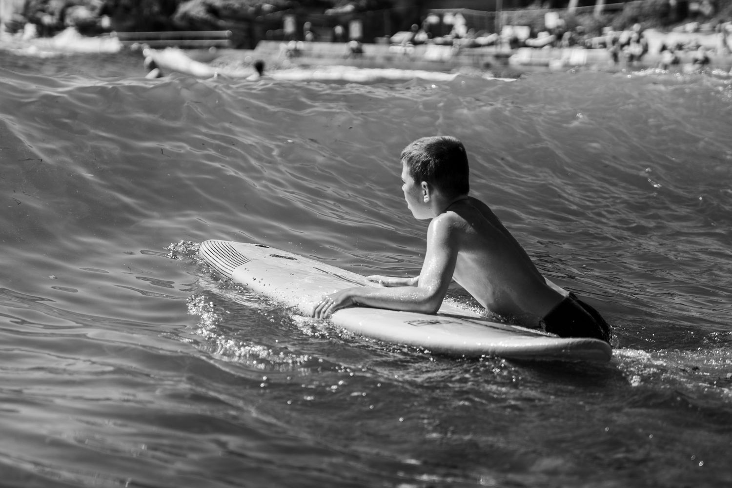 Black and white photo of a young boy lying on a surfboard paddling through ocean waves.
