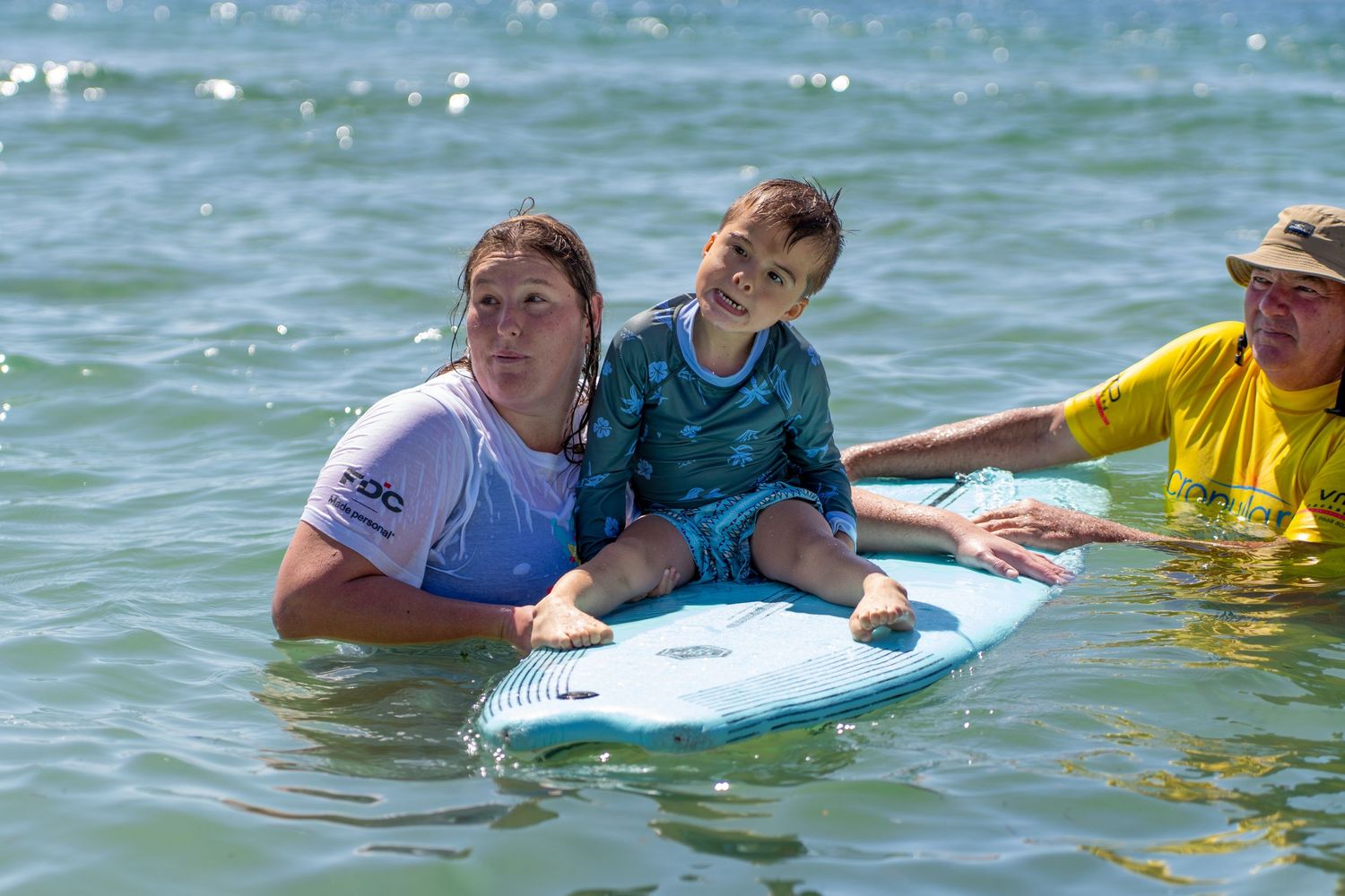 Two surf instructors helping a young child balance on a surfboard in calm ocean water.