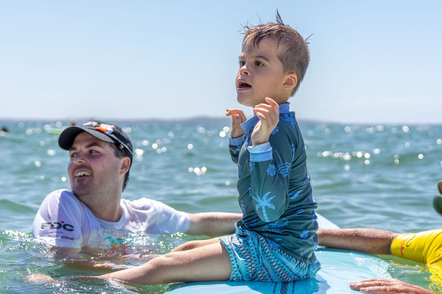 Male surf instructor supporting a young child standing on a surfboard in the ocean.