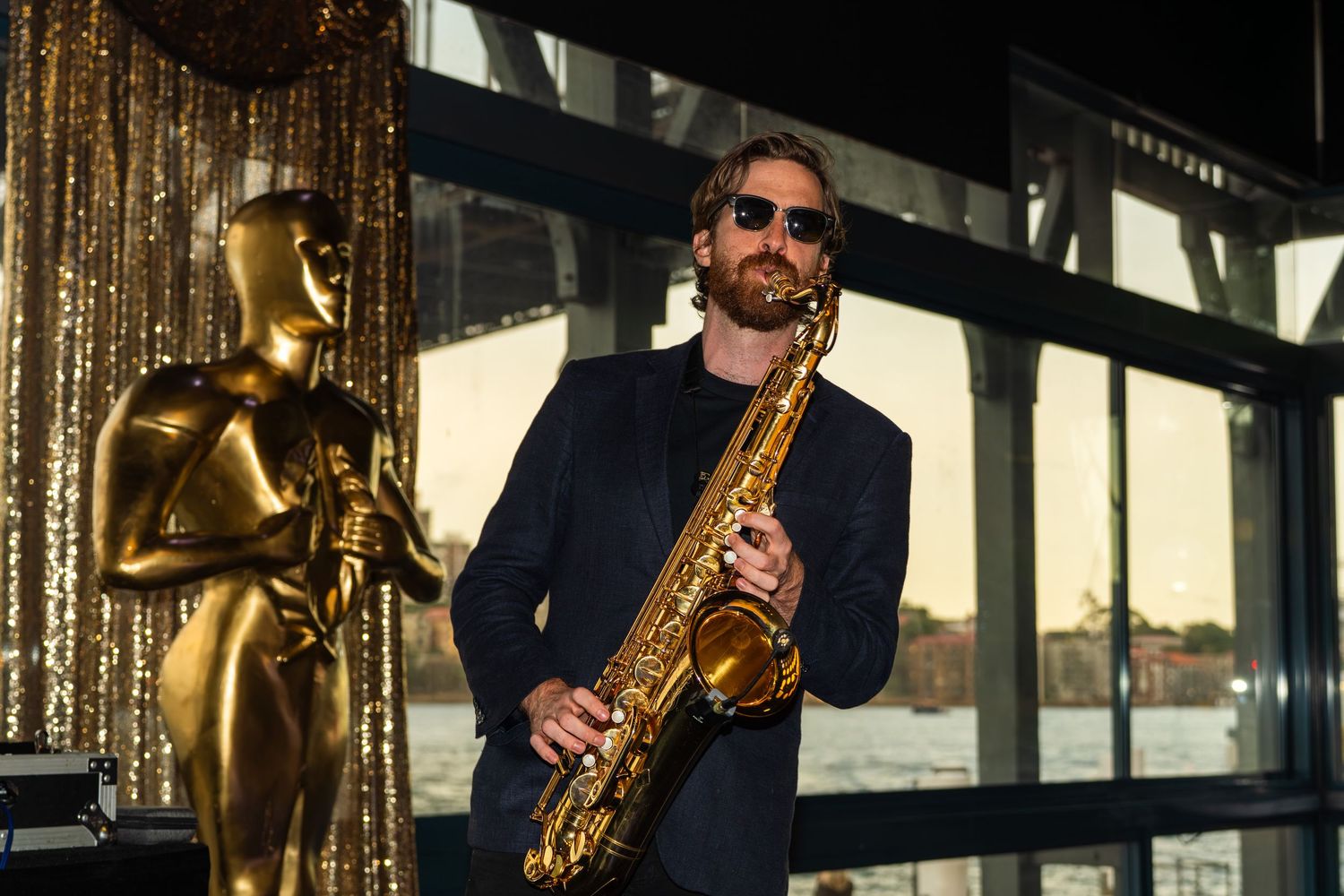 Saxophone player posing next to a gold Oscar statue at a glamorous event.