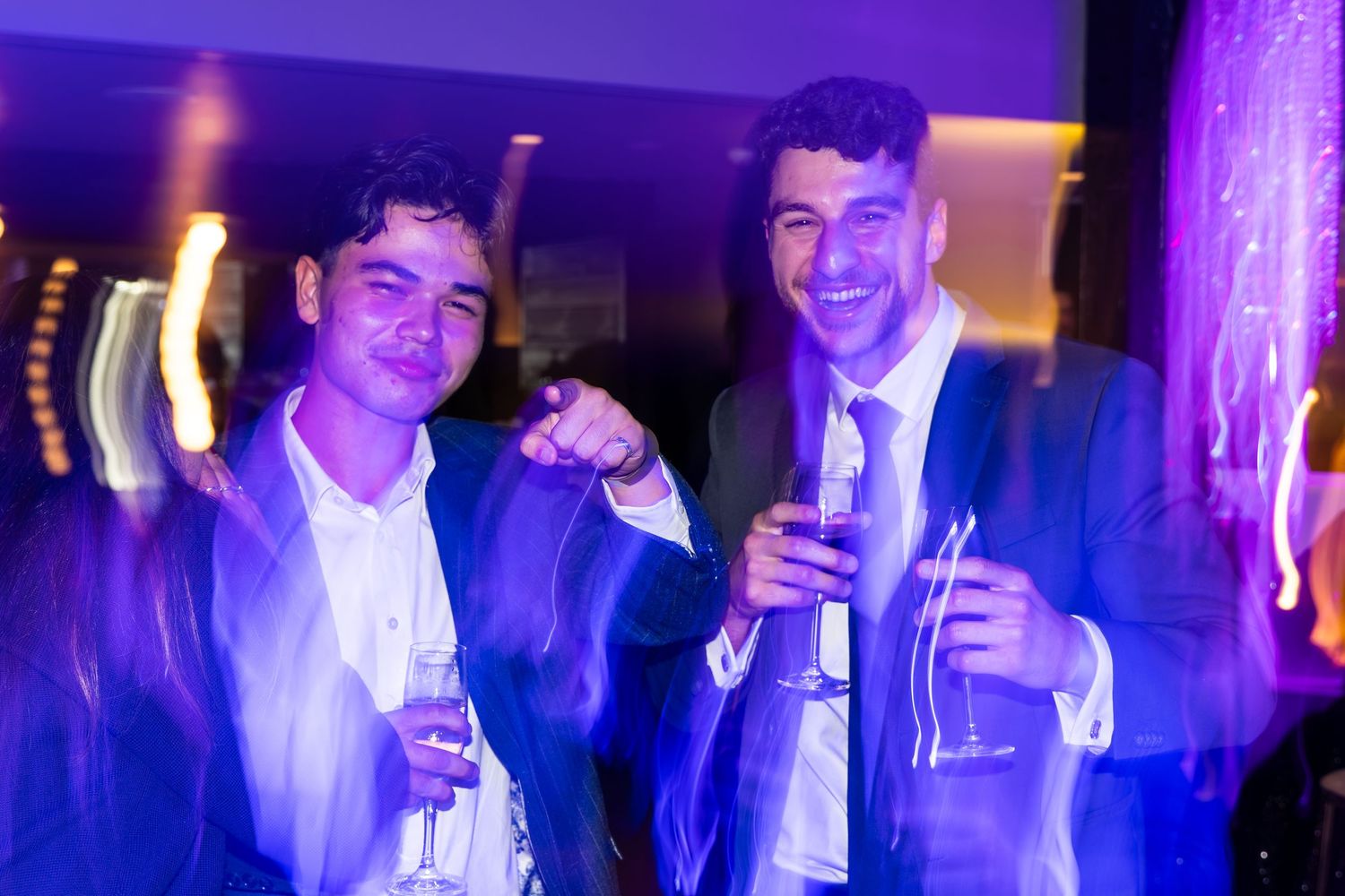 Two men in suits smiling and holding drinks at a blue-lit party.