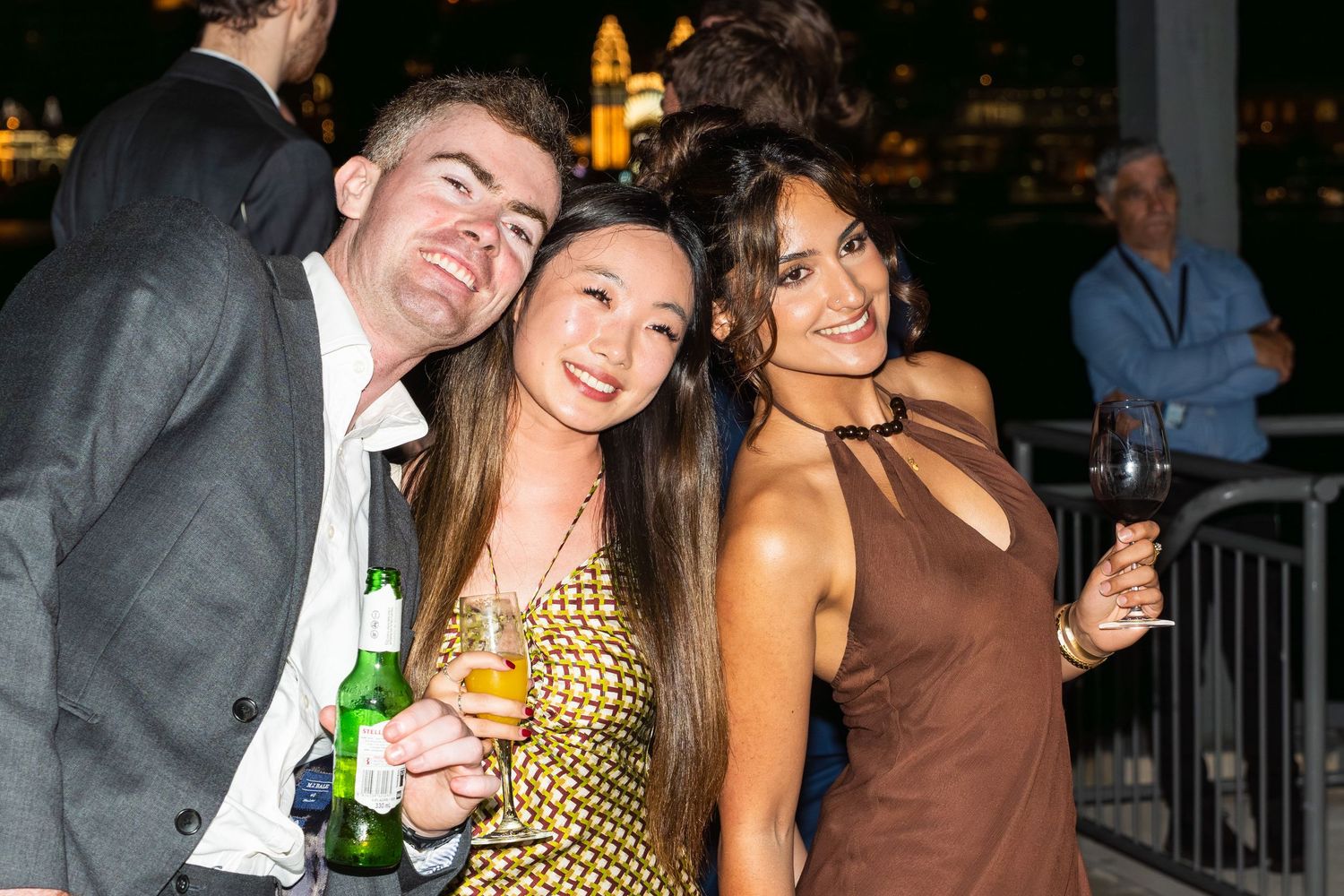 Three friends smiling and holding drinks at an outdoor evening event.