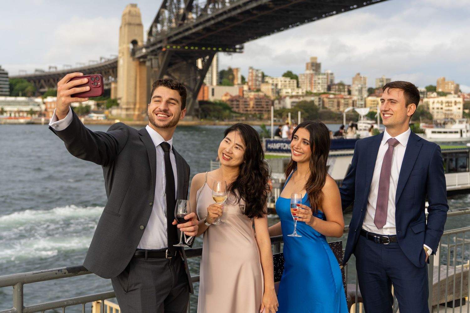Four formally dressed young adults take a selfie with Sydney Harbour Bridge in background.