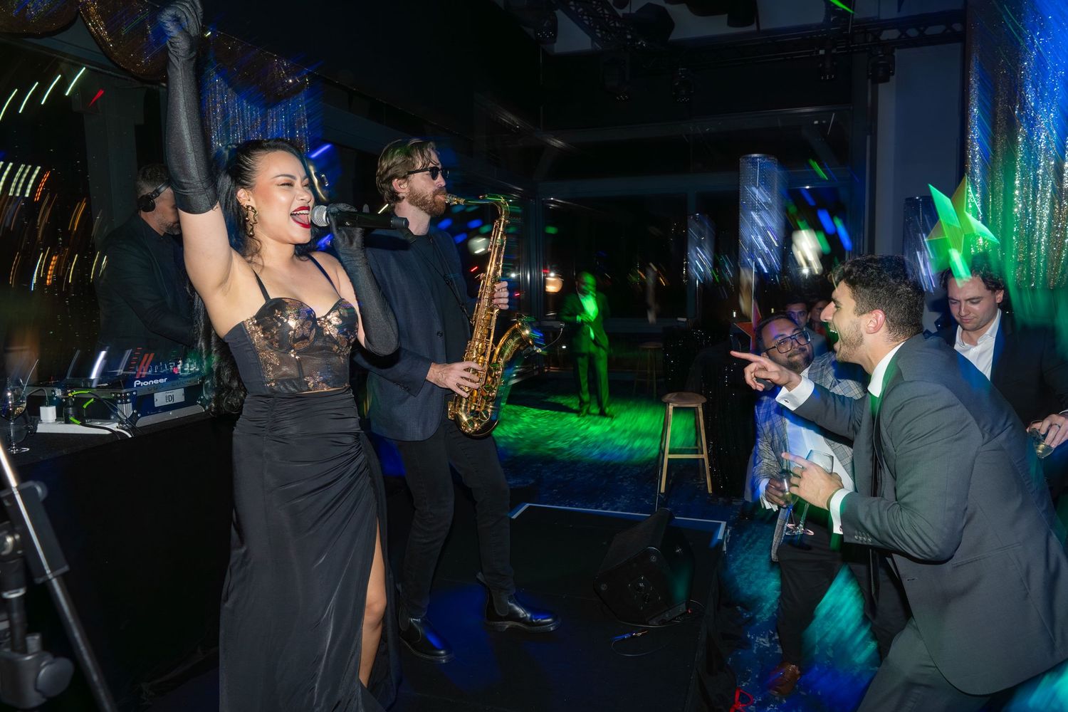 A performer in a black dress sings while a saxophonist plays at a lively nightclub event.