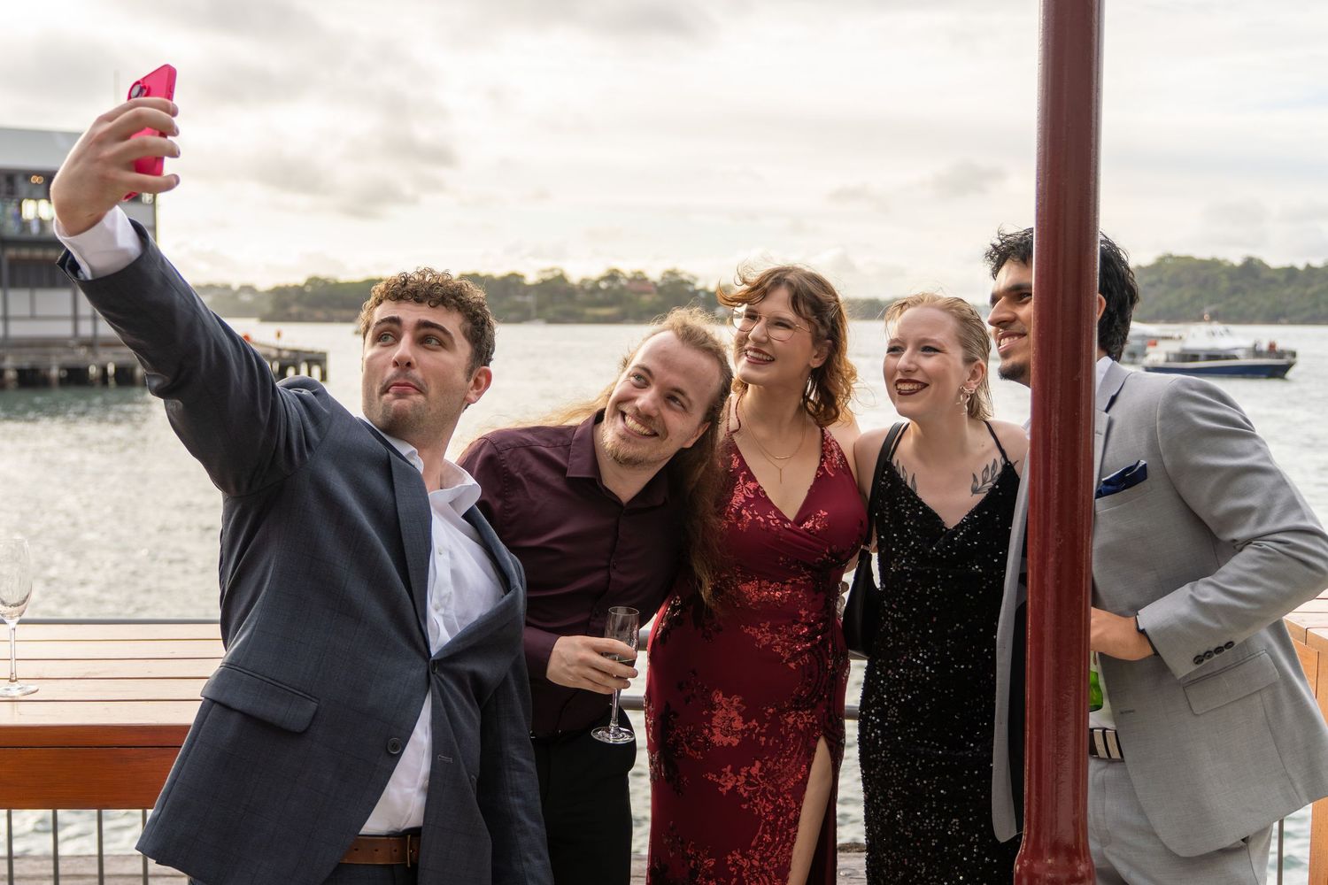 Group of five young adults in formal wear taking a selfie on a waterfront terrace in Sydney.