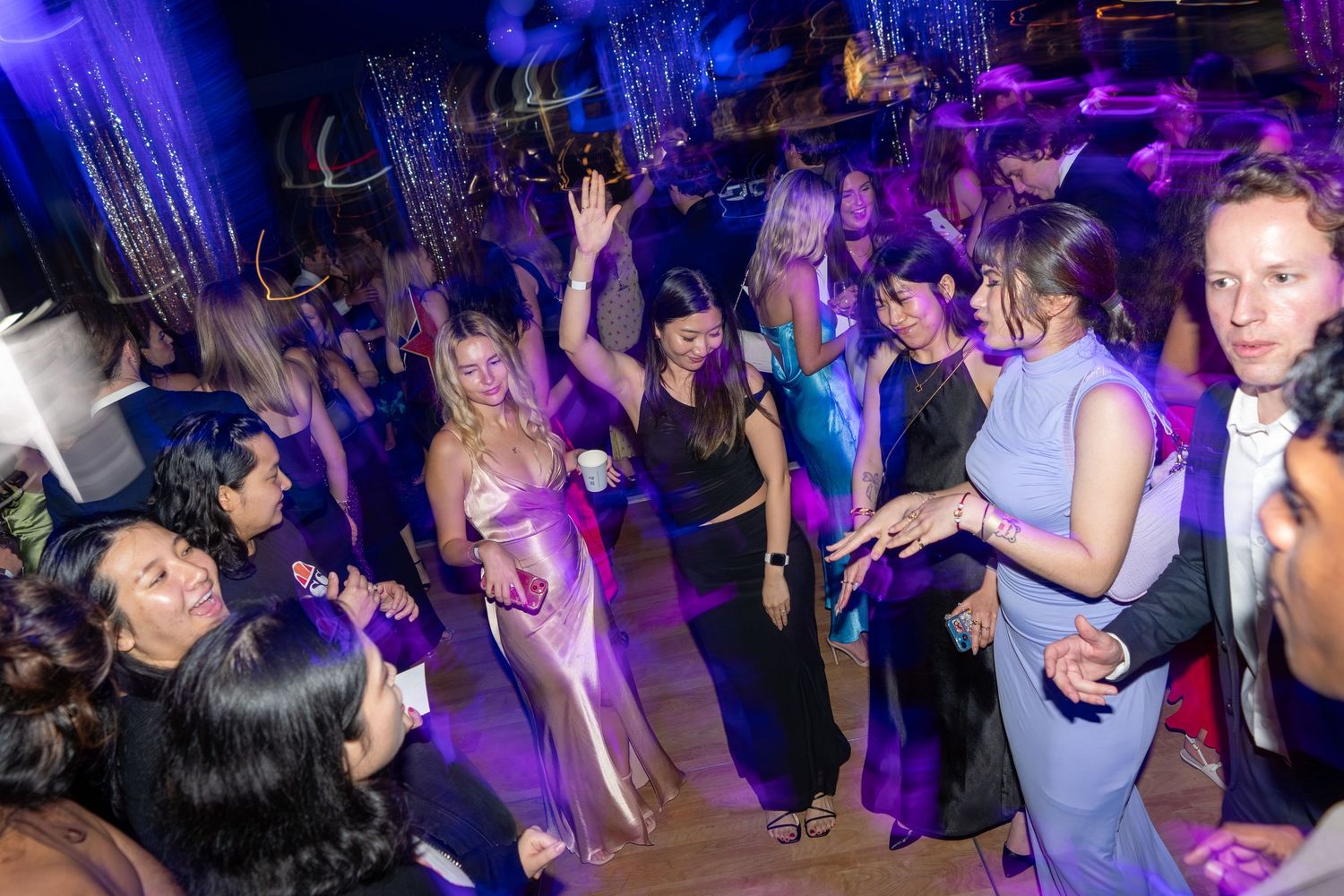 A large group of elegantly dressed guests dance together under purple lights at a gala.