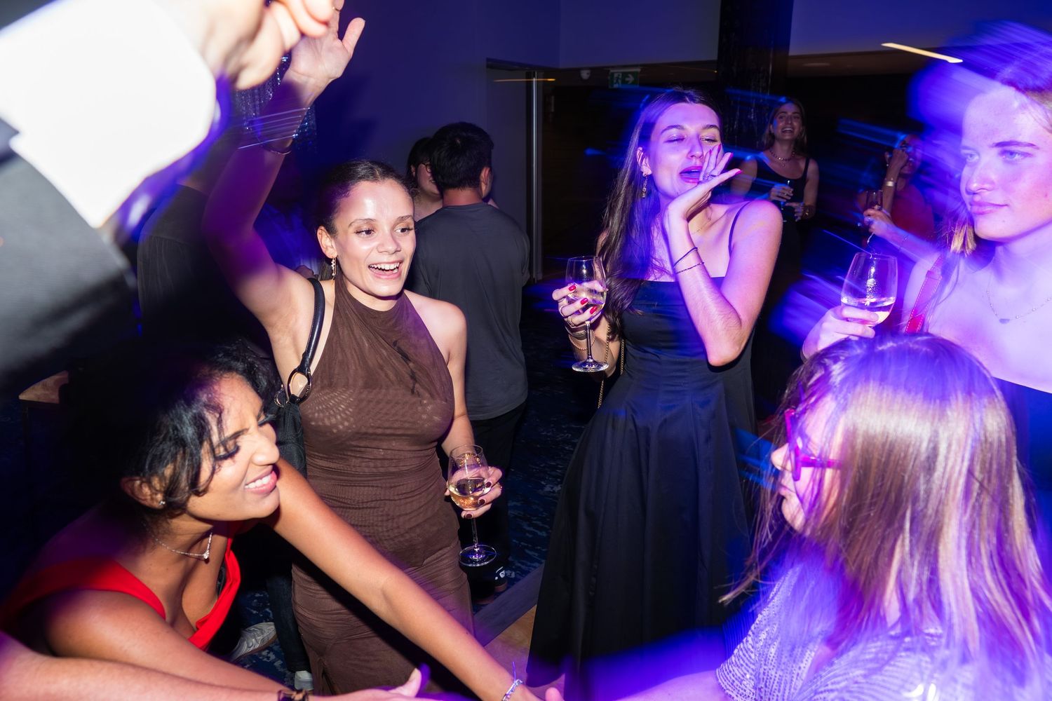 Young women in formal attire laugh and dance together under blue party lights.