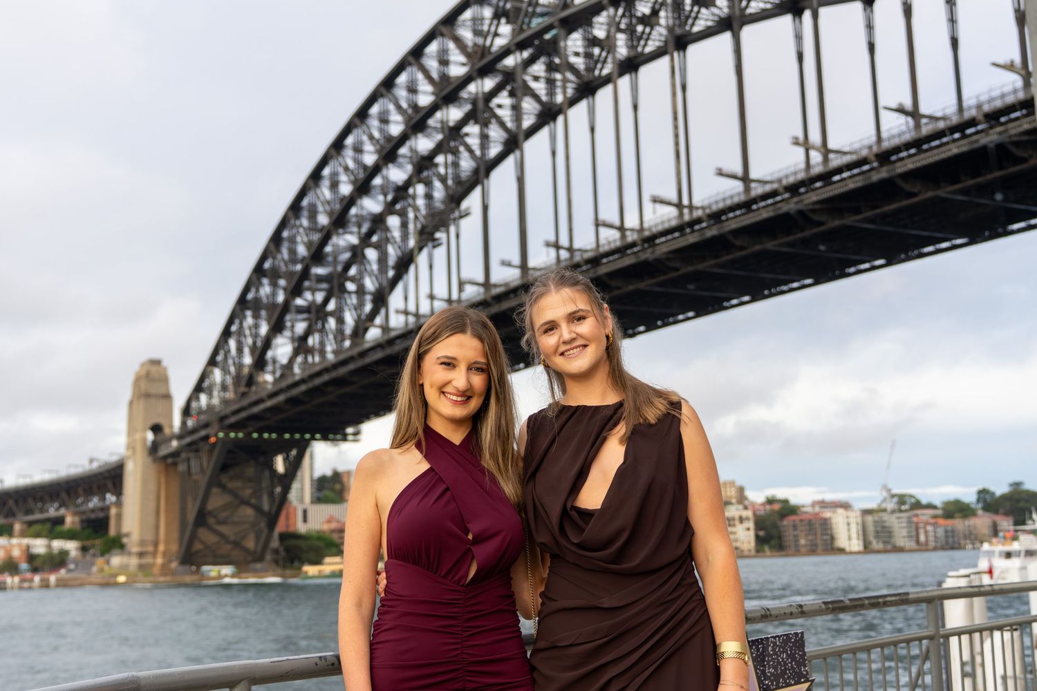 Two women in elegant dresses posing in front of the iconic Sydney Harbour Bridge.