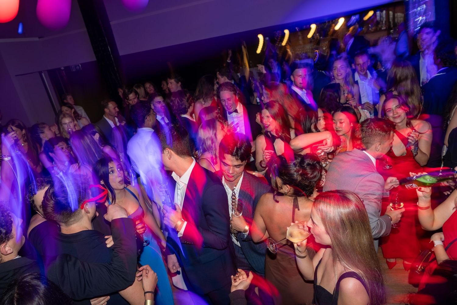 Crowded nightclub dance floor with people dancing under colorful purple and pink lights at a lively party event.