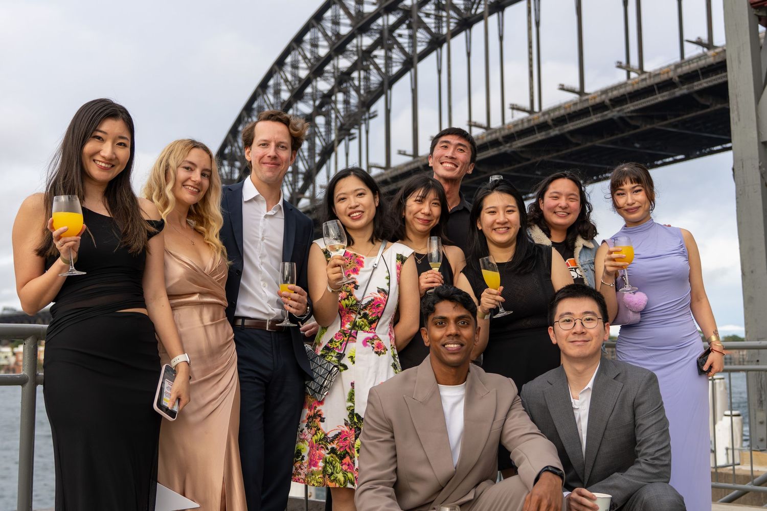 Diverse group of formally dressed friends posing with drinks at Sydney Harbour Bridge event.