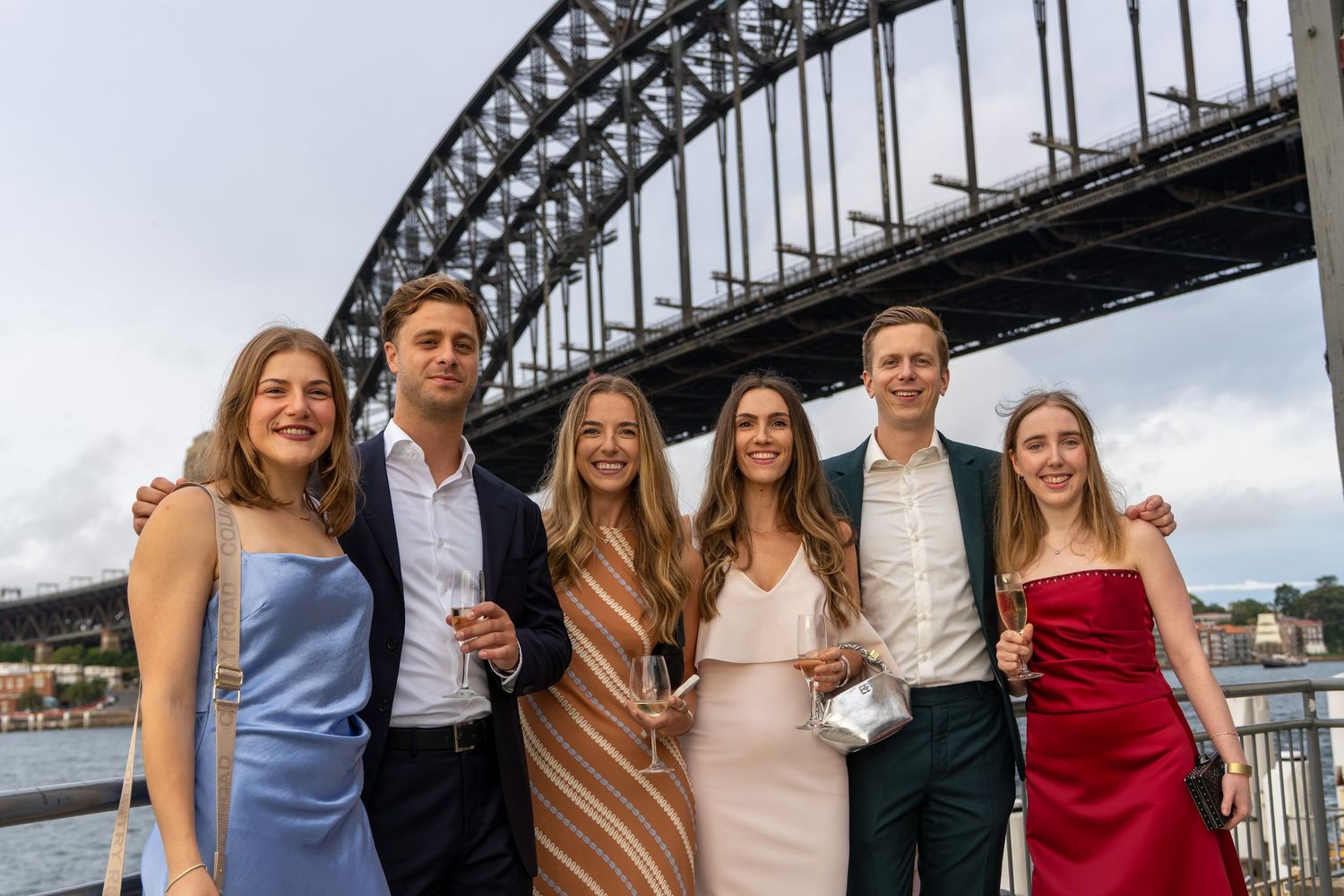 Six young adults in formal attire smiling together with Sydney Harbour Bridge backdrop.