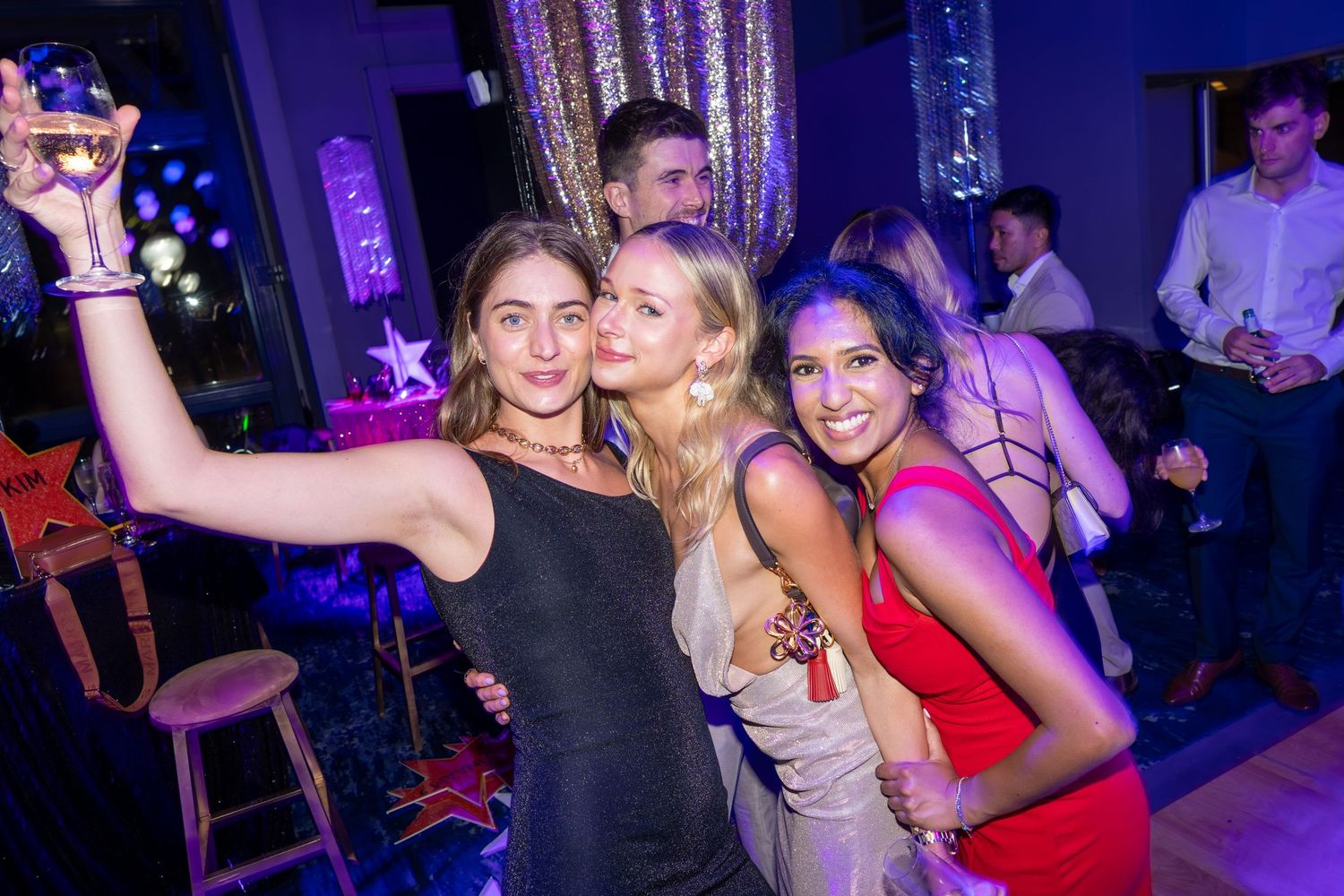Three women smiling and taking a selfie with a cocktail at a nightclub party with blue ambient lighting.