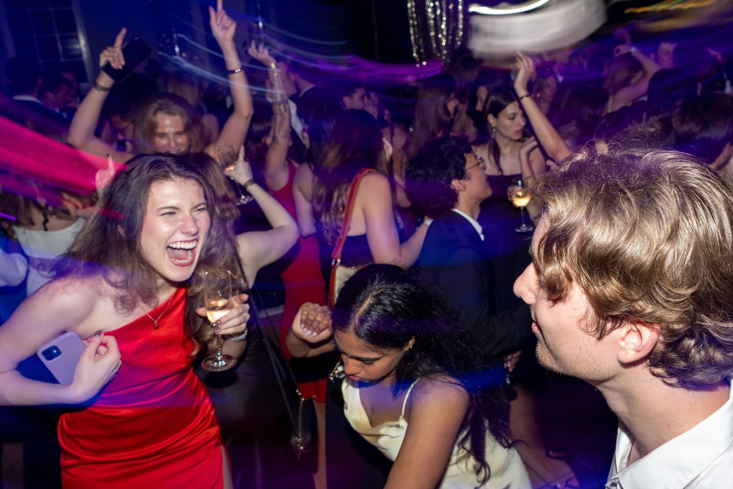 Young woman in red dress laughing and dancing at a crowded nightclub party with blue and purple lights.