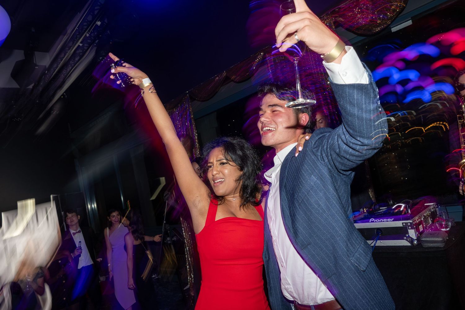 Couple dancing and raising glasses at a vibrant nightclub party with colorful lighting in the background.