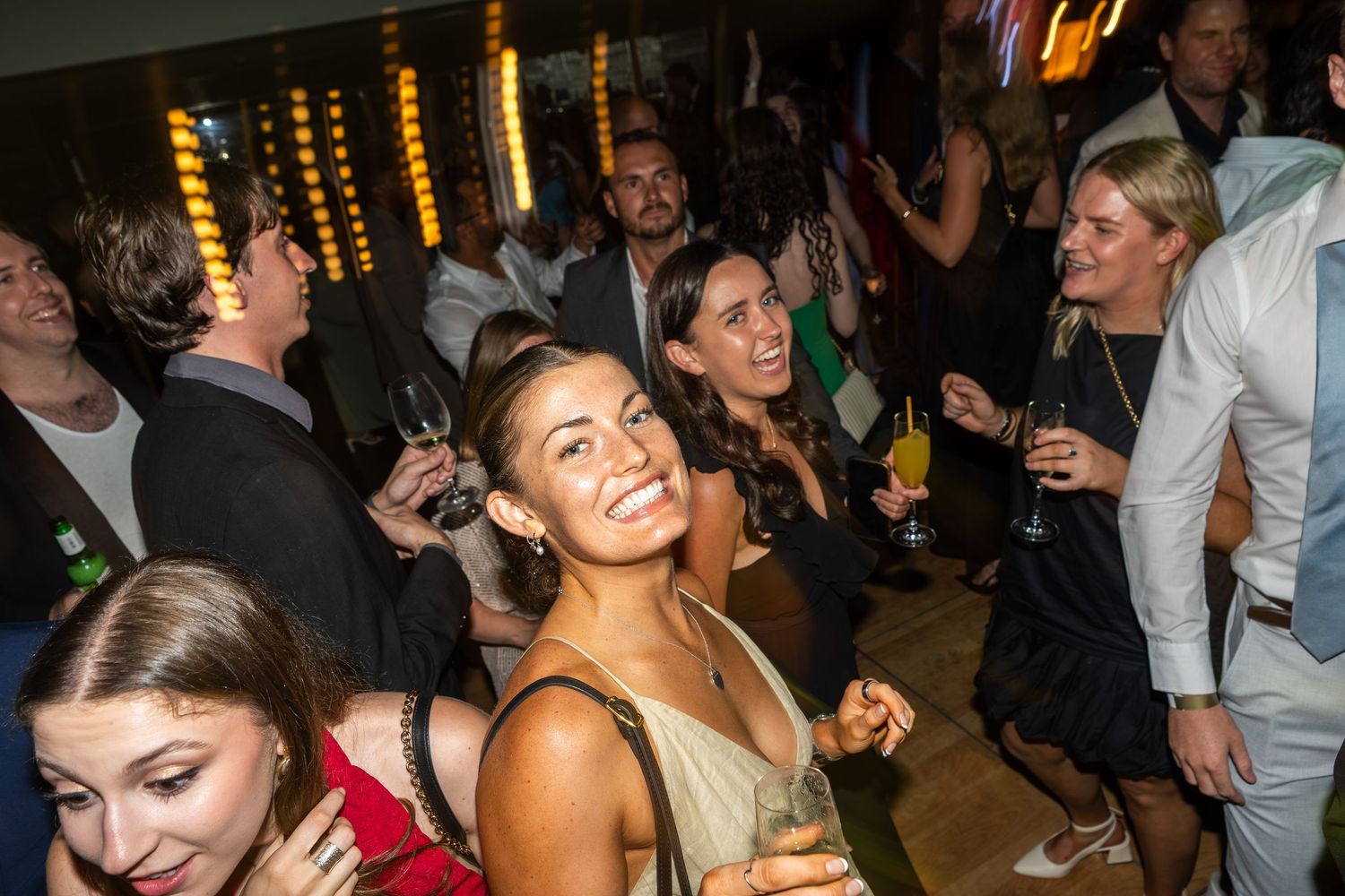 Group of elegantly dressed people socializing and dancing at an upscale indoor party event with warm lighting.