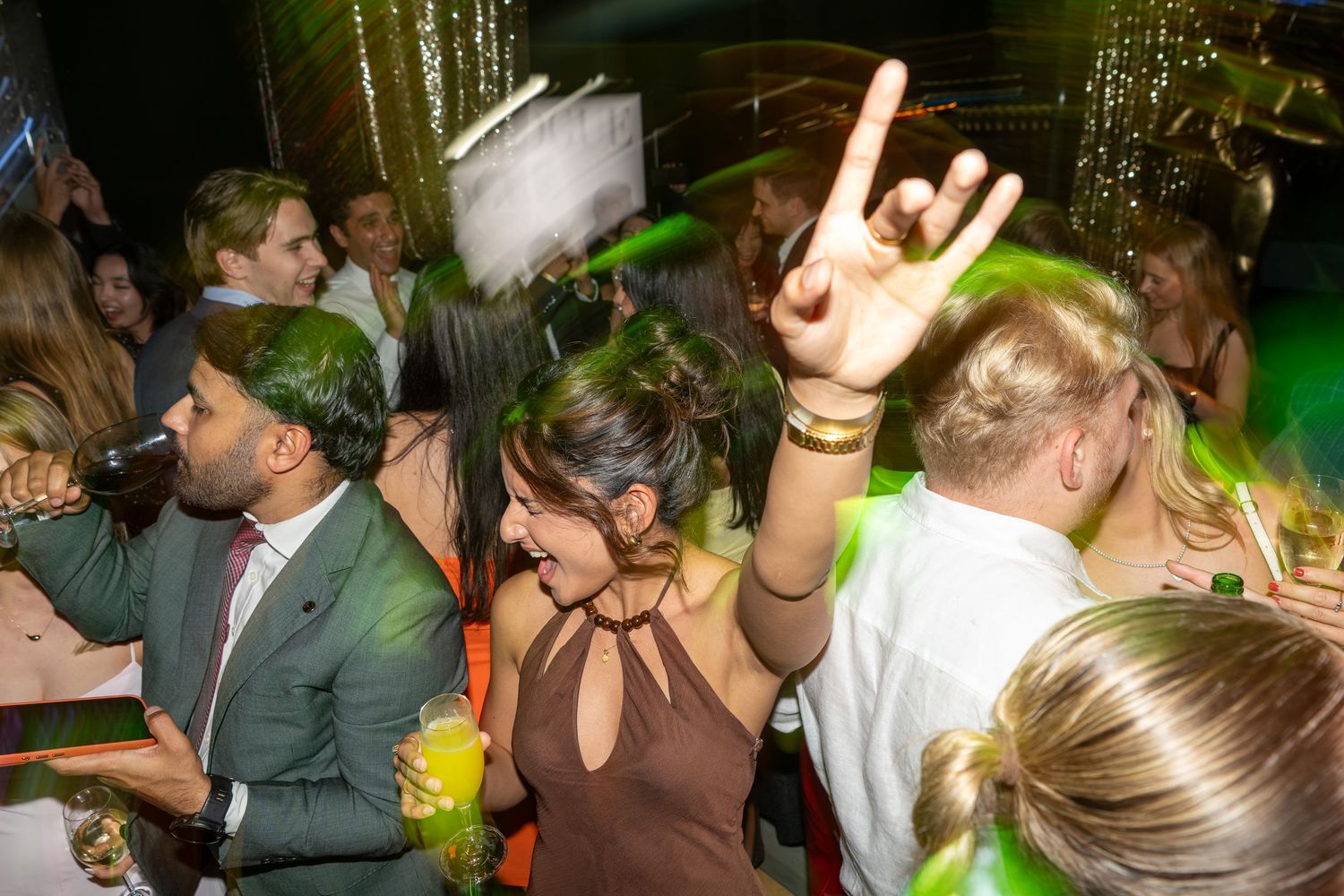 Woman making rock hand gesture while dancing at a lively party with green-lit decorations and crowd.