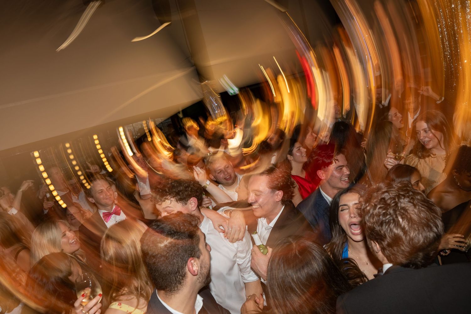 Motion-blurred crowd of party guests dancing energetically under warm golden lights at a festive event.