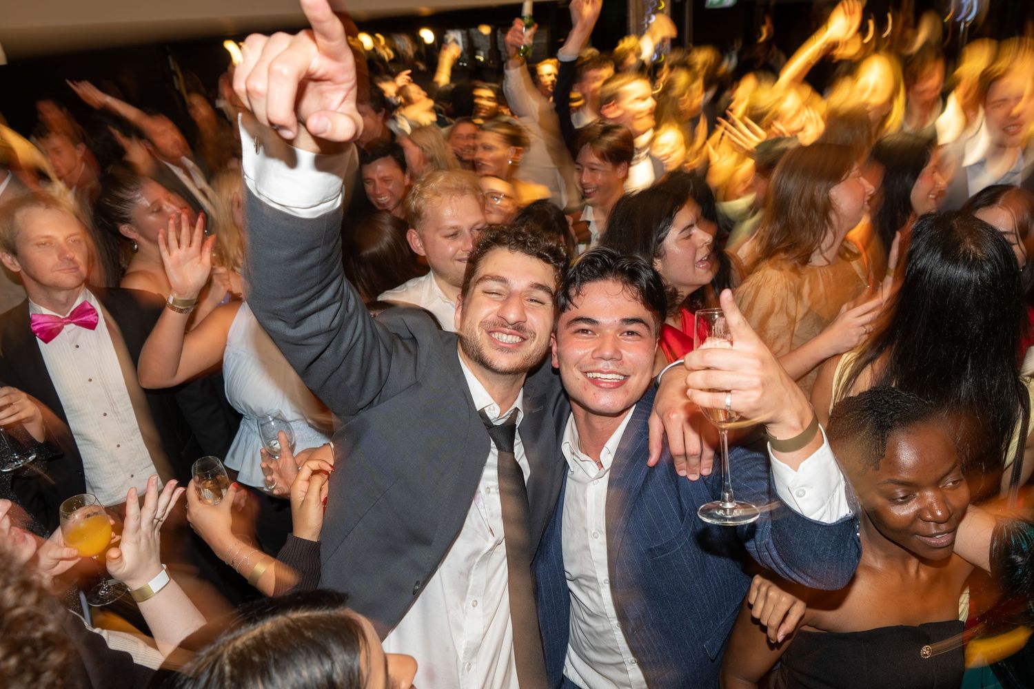 Two young men smiling and celebrating with drinks raised at a lively crowded party event.