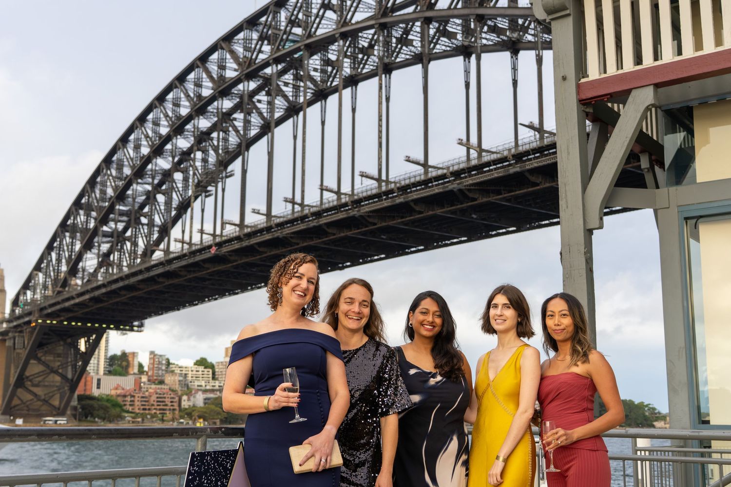 Five women in elegant dresses posing with drinks at Sydney Harbour Bridge waterfront.
