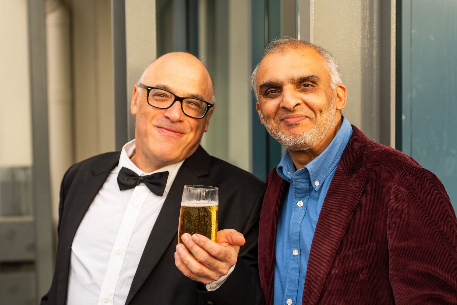 Two men in formal attire smiling and holding drinks at an indoor event.