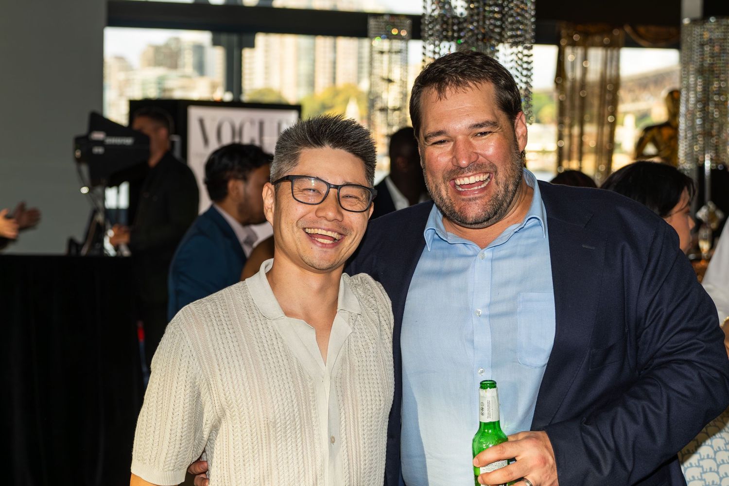 Two men smiling together at a Vogue-themed end of year party holding drinks.