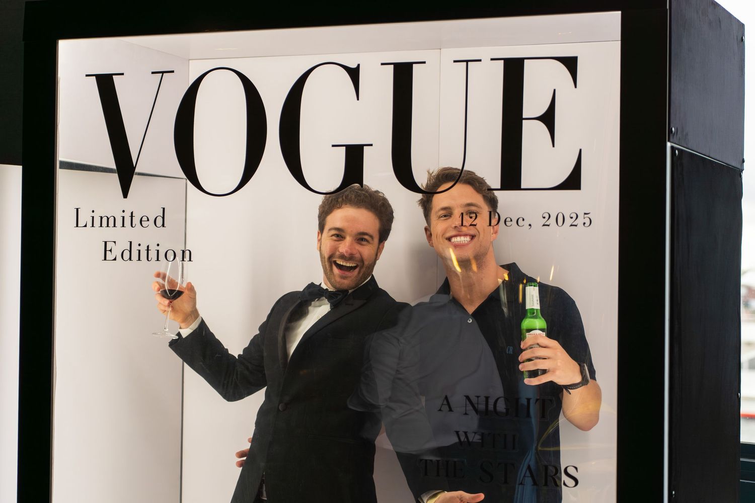Two men posing with drinks in front of a Vogue Limited Edition photo booth.