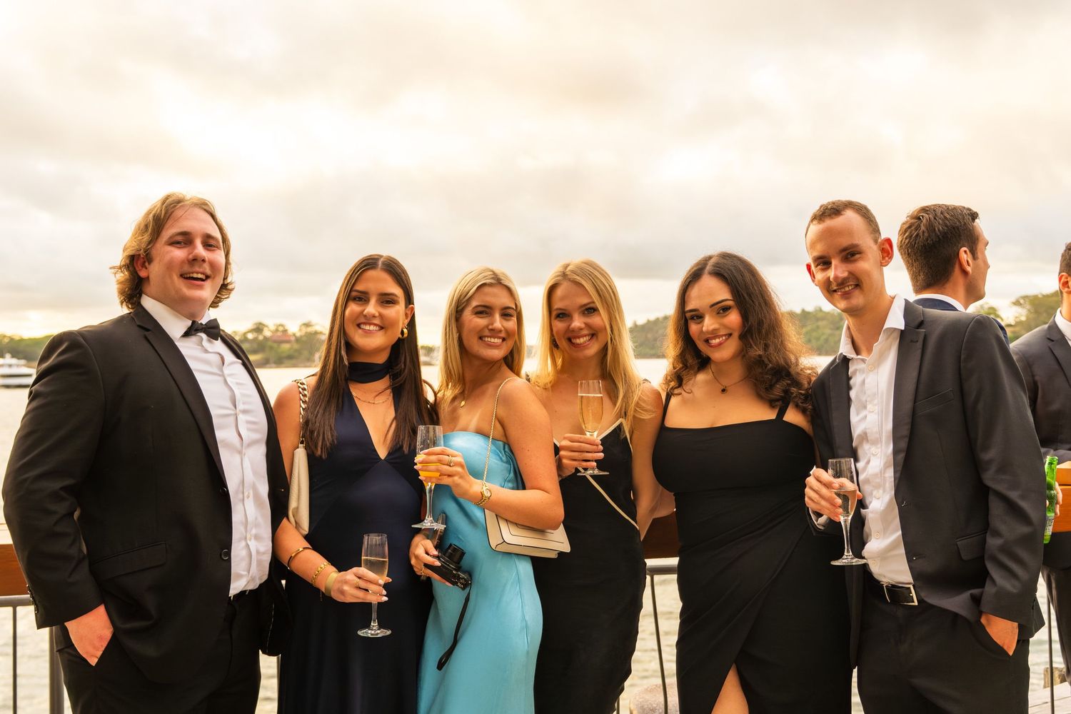 Six formally dressed guests smiling with champagne at a waterfront evening party.