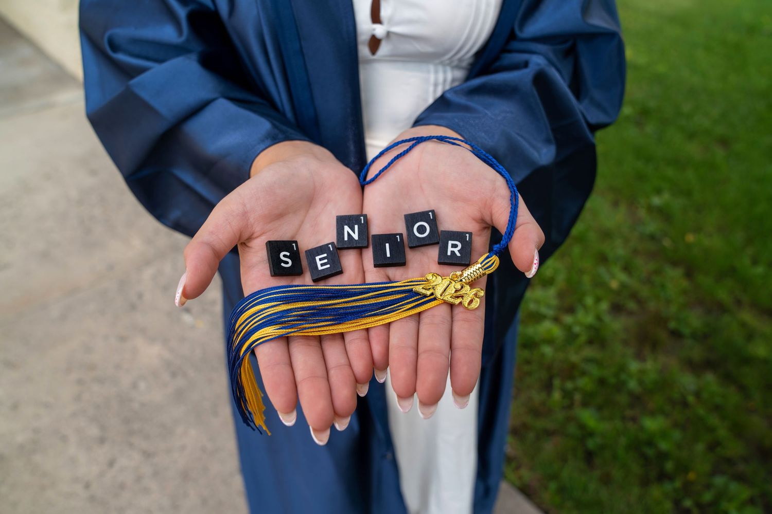 Graduate in blue cap and gown holds Scrabble tiles spelling SENIOR with gold tassel in hands.