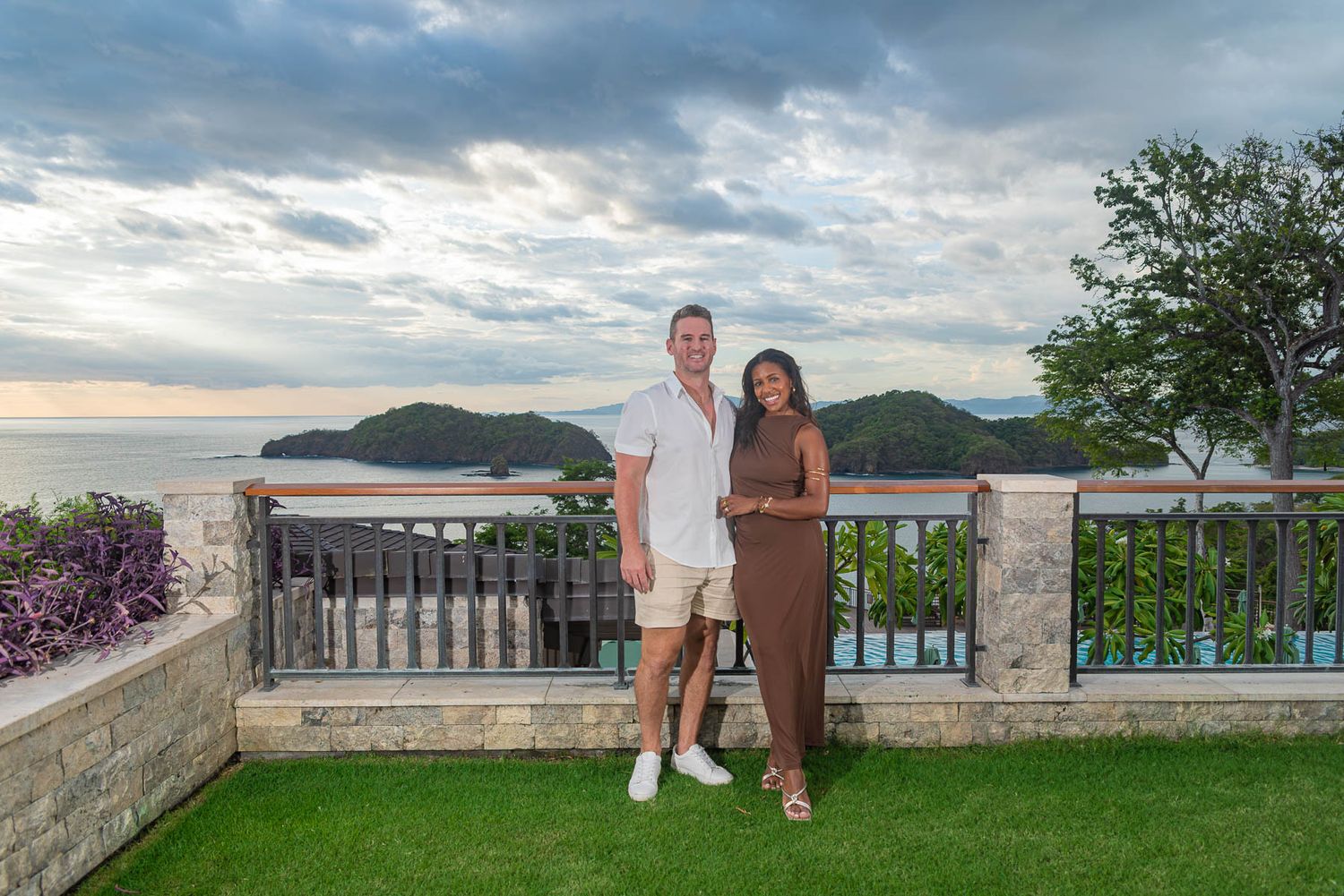 Couple posing on a terrace with ocean and mountain views at sunset, lush greenery and dramatic cloudy sky.