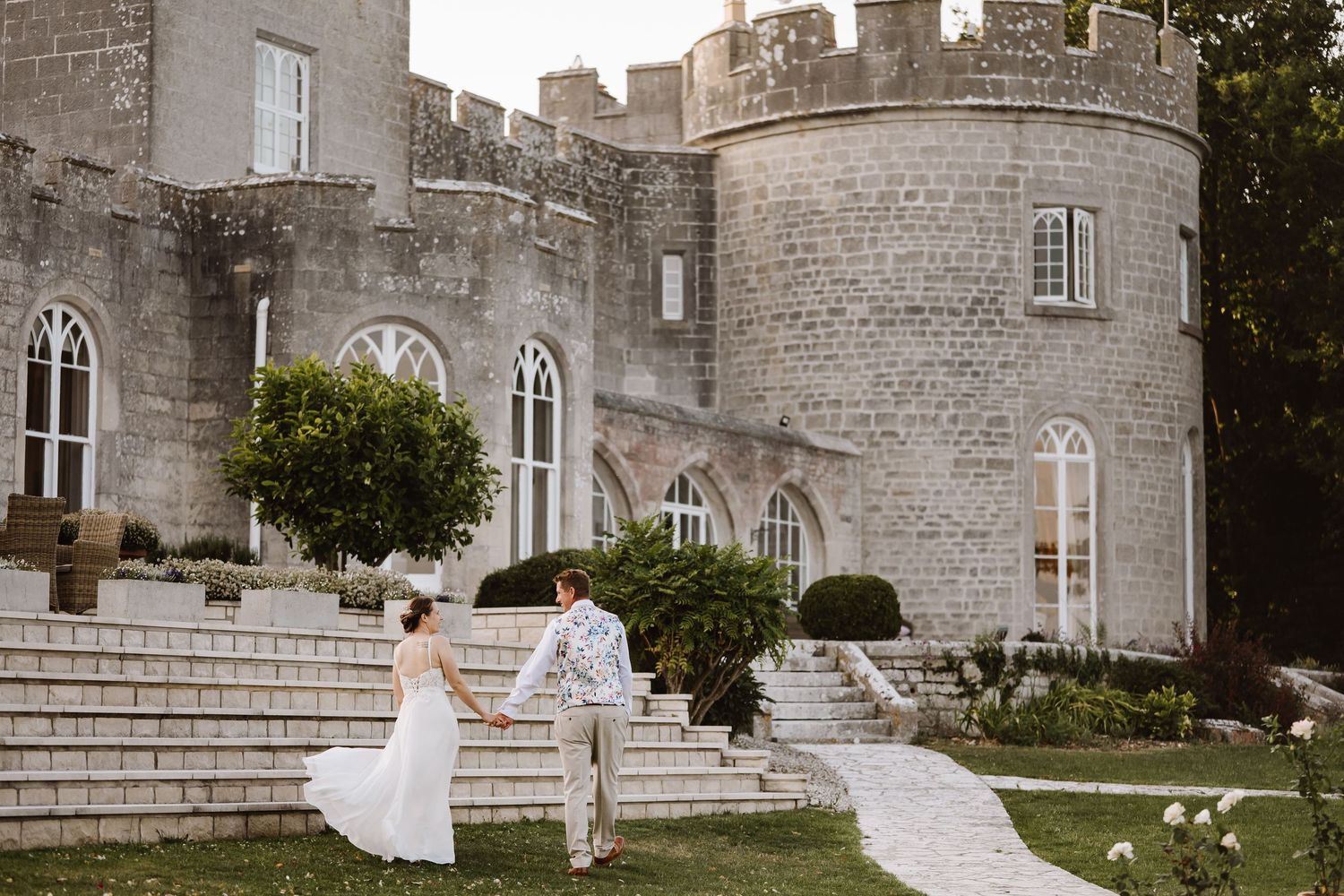 Bride and groom walk hand in hand toward a stunning stone castle wedding venue with arched windows and lush gardens.