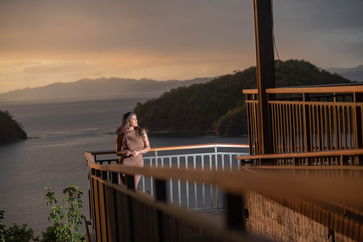 Woman standing on a scenic overlook deck at sunset with ocean and mountain views in the background.