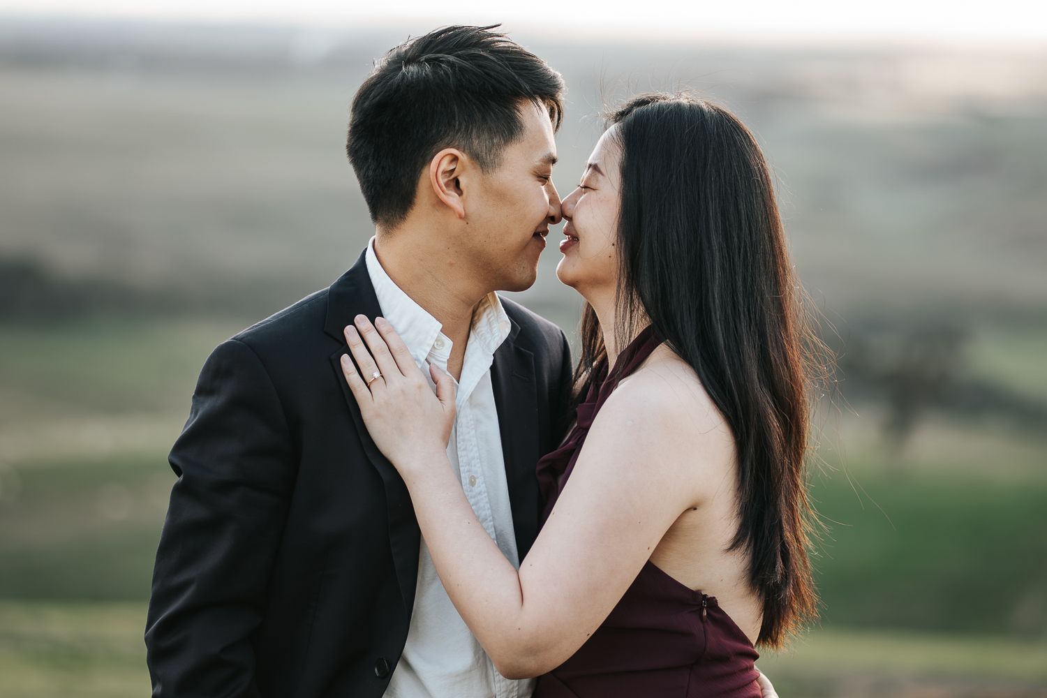 Couple sharing a romantic kiss while embracing on a green hillside with scenic rolling countryside views at sunset.