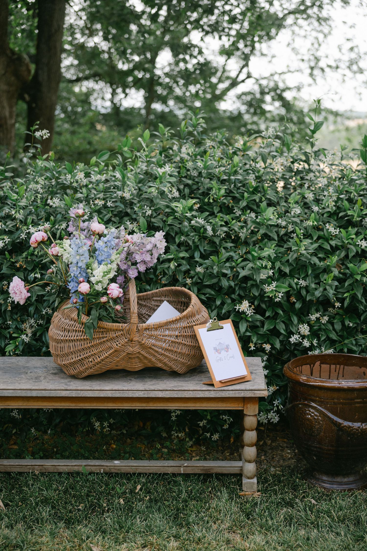 Wicker basket with pink and blue flowers on a wooden bench beside a clipboard, set against lush green garden shrubs.