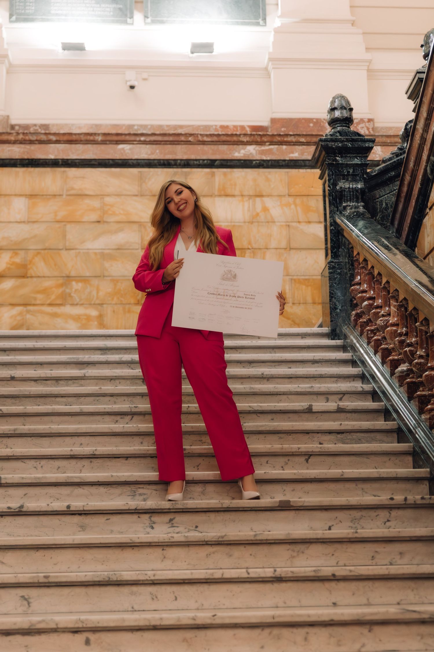 Woman in bright pink suit poses on grand staircase holding a large framed certificate.