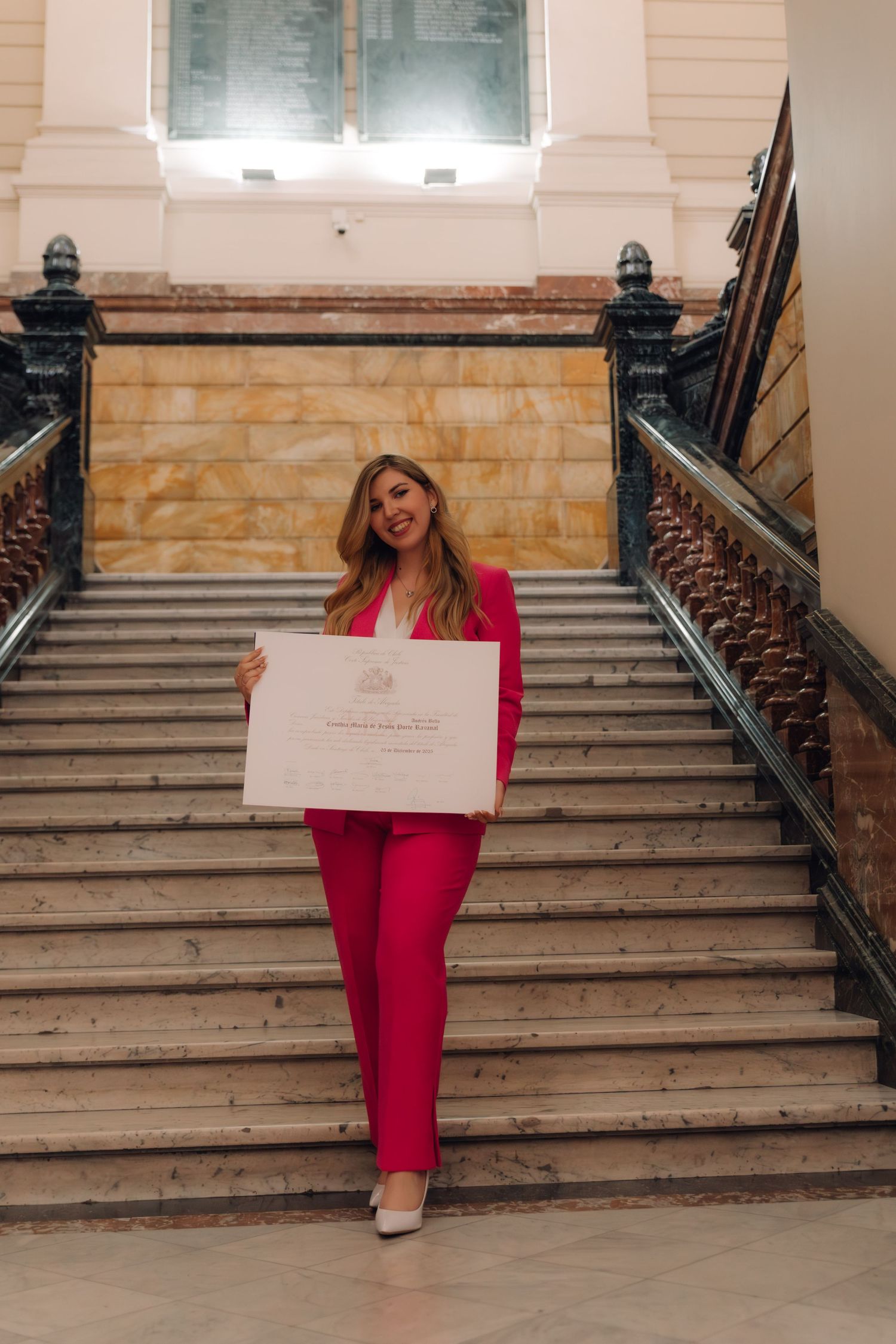 Elegant woman in pink pantsuit stands on marble staircase displaying a graduation certificate.