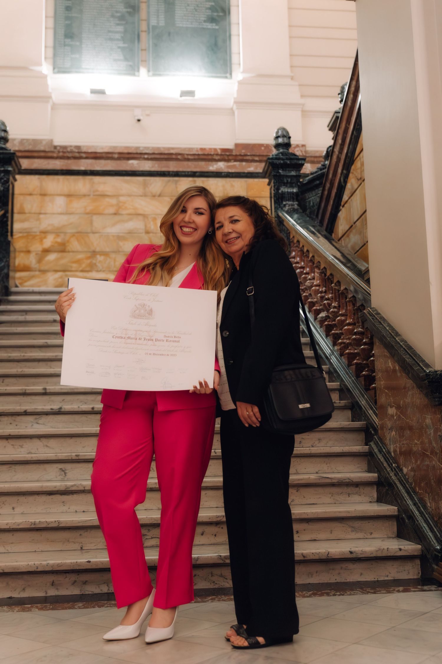 Two women posing with a diploma on a grand staircase, one in pink suit.