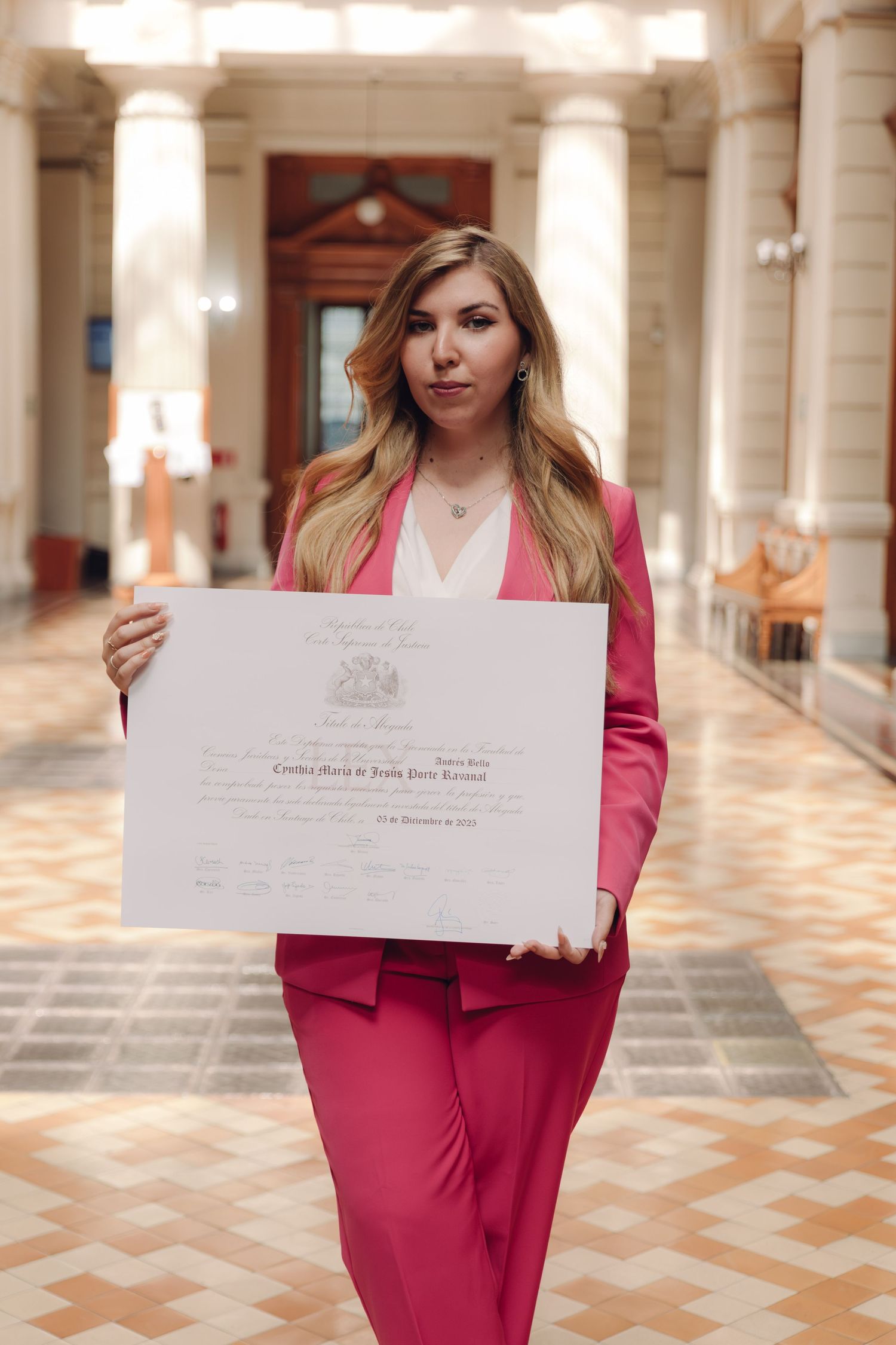 Woman in pink suit holding a diploma certificate in an elegant columned hallway.