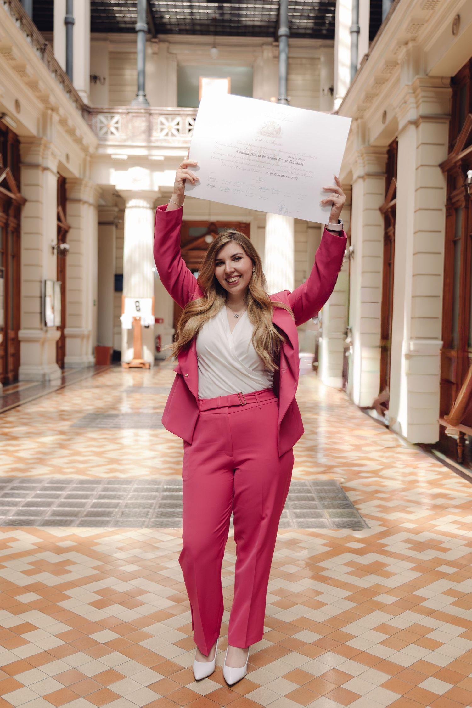 Joyful woman in pink suit raising diploma overhead in a classical building hallway.