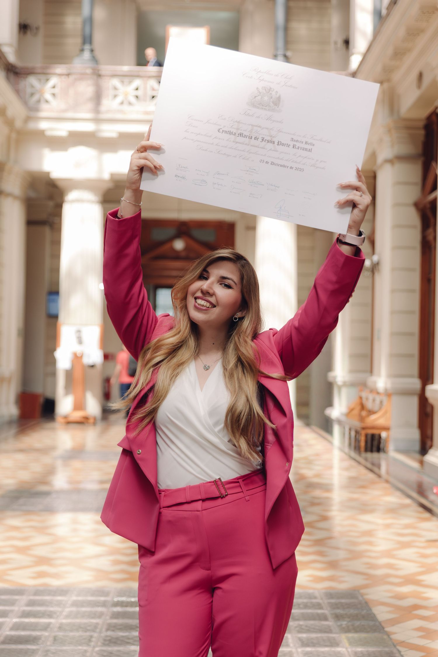 Excited graduate in pink suit lifting diploma certificate high in ornate hall.