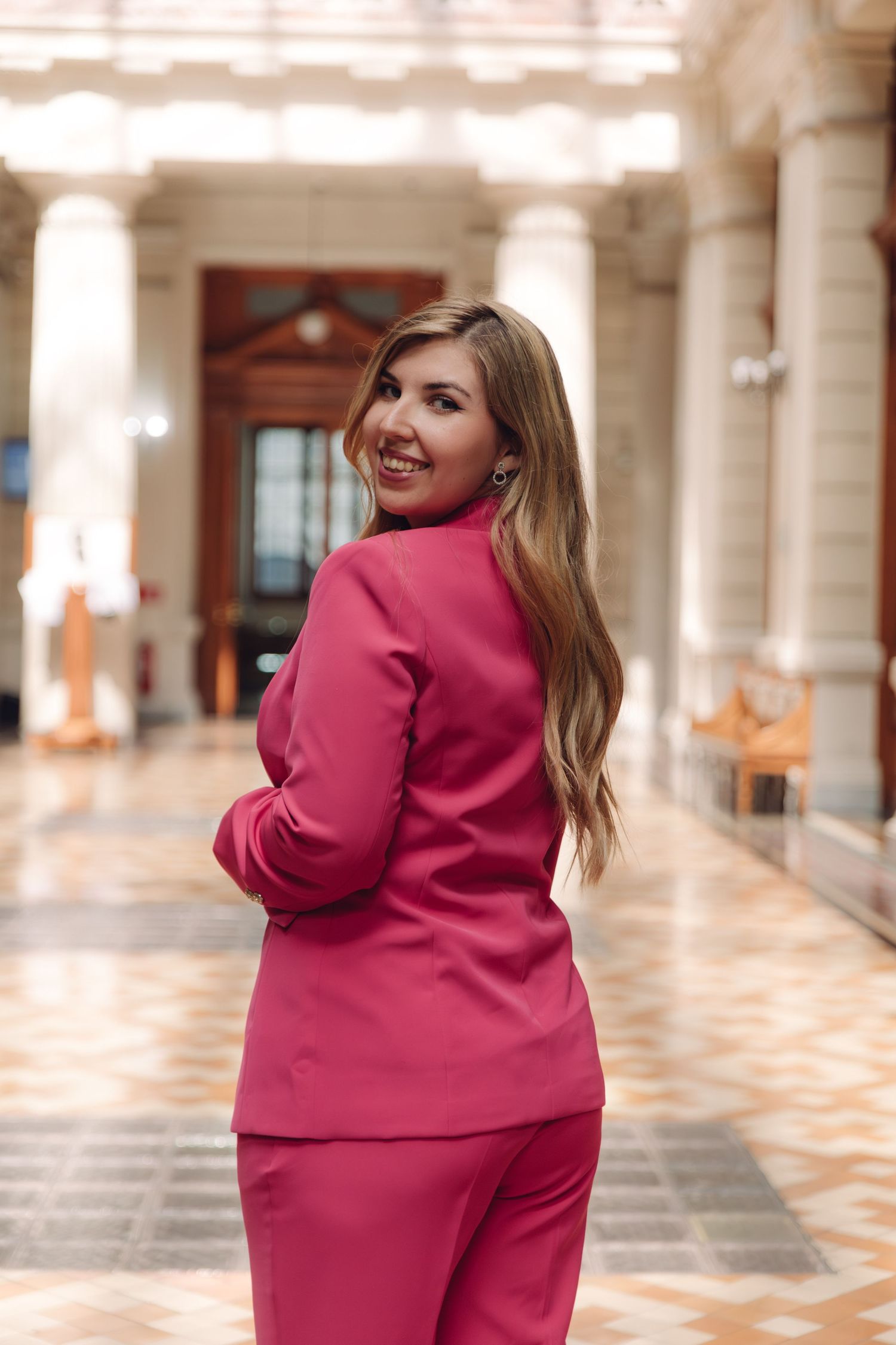 Stylish woman in pink suit smiling over shoulder in grand columned corridor.