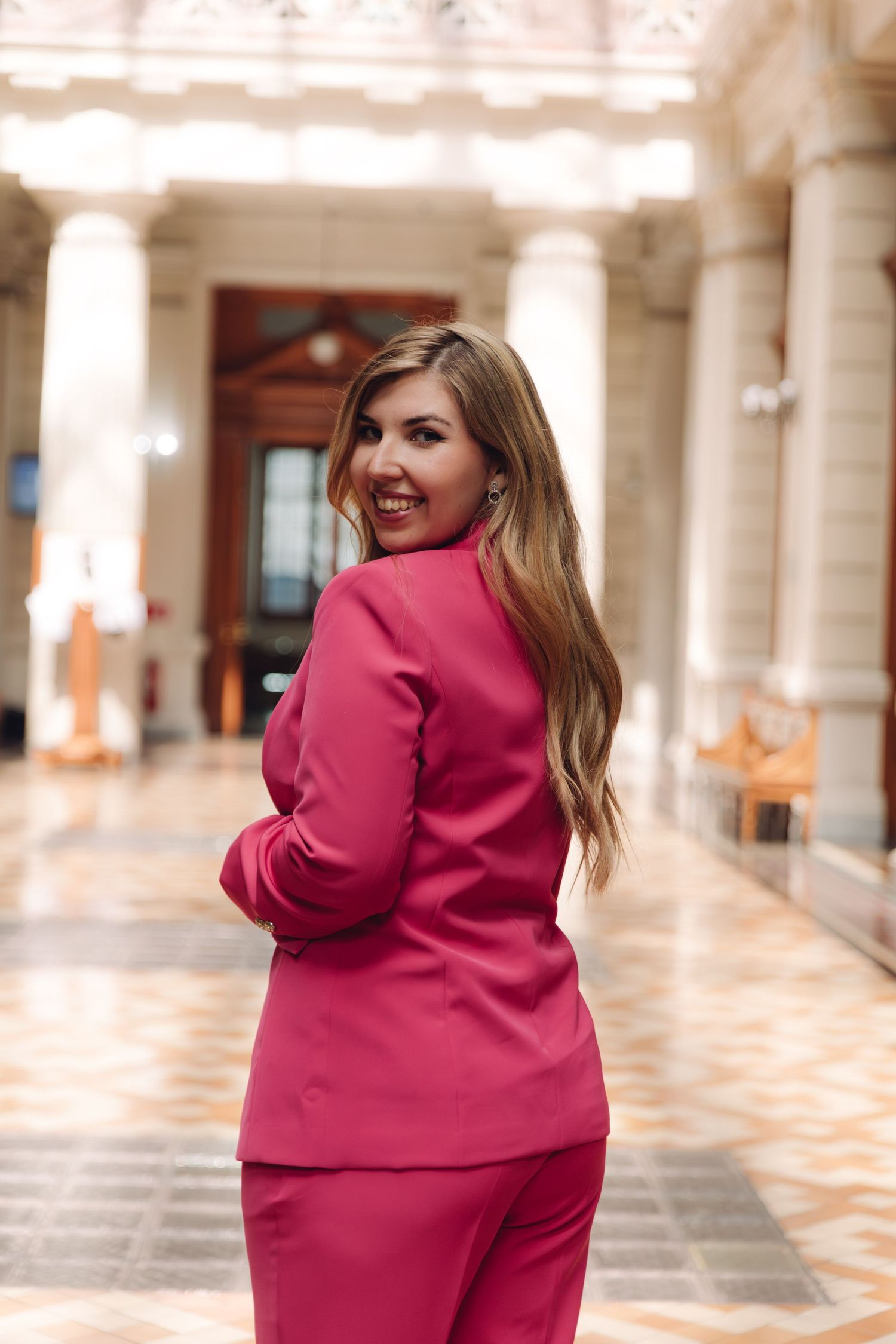 Elegant woman in fuchsia pantsuit turning to smile in a classical building.