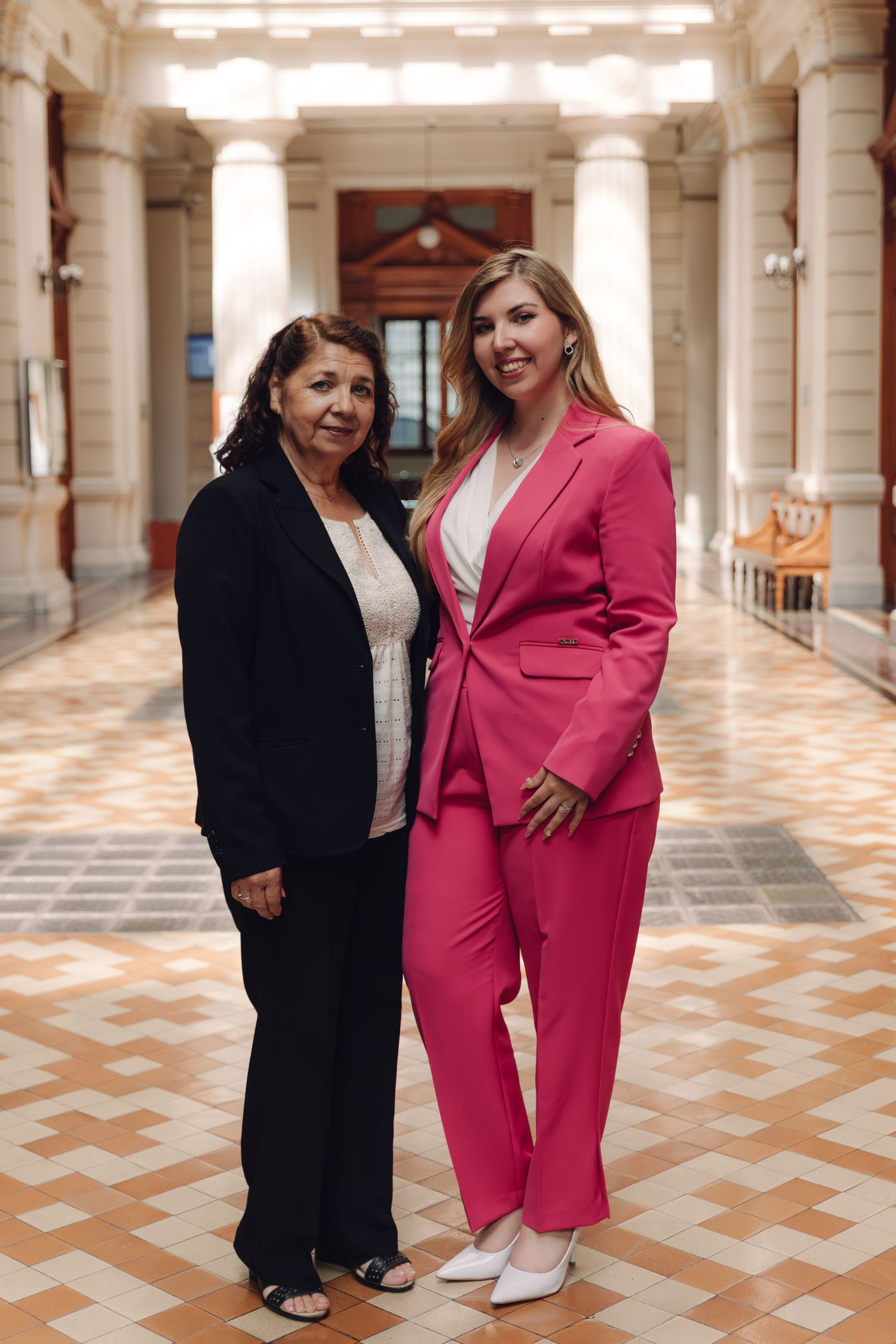 Two women posing together in elegant hallway, one wearing a bright pink suit.