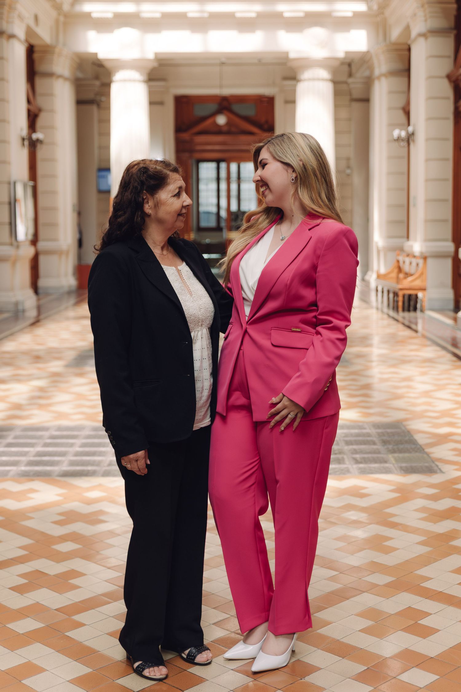 Two women laughing together in a grand columned hall, one in pink suit.