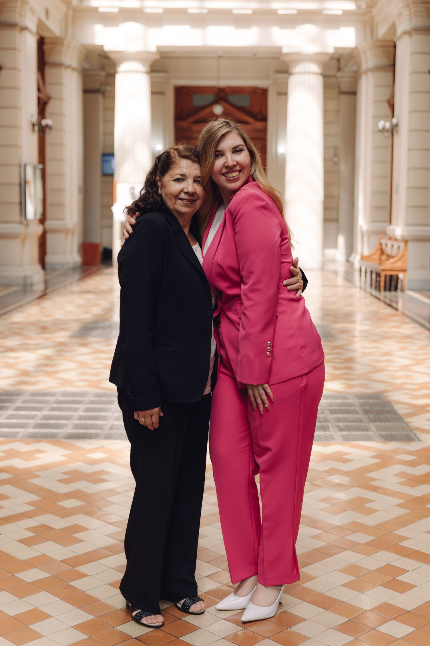 Two women in black and pink suits embrace and smile in an elegant columned hallway.