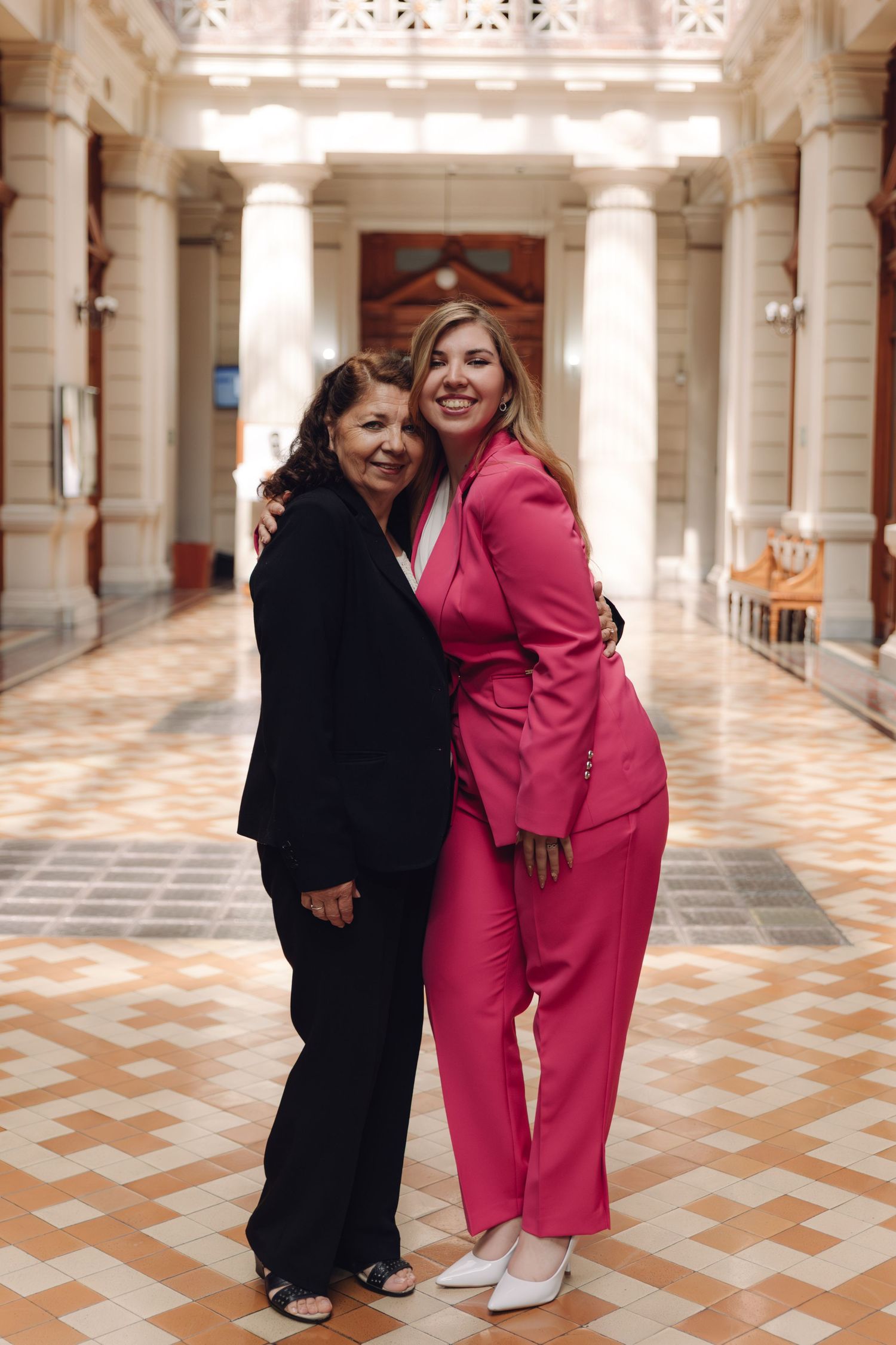 Two women in black and pink pantsuits pose together smiling in a grand architectural corridor.