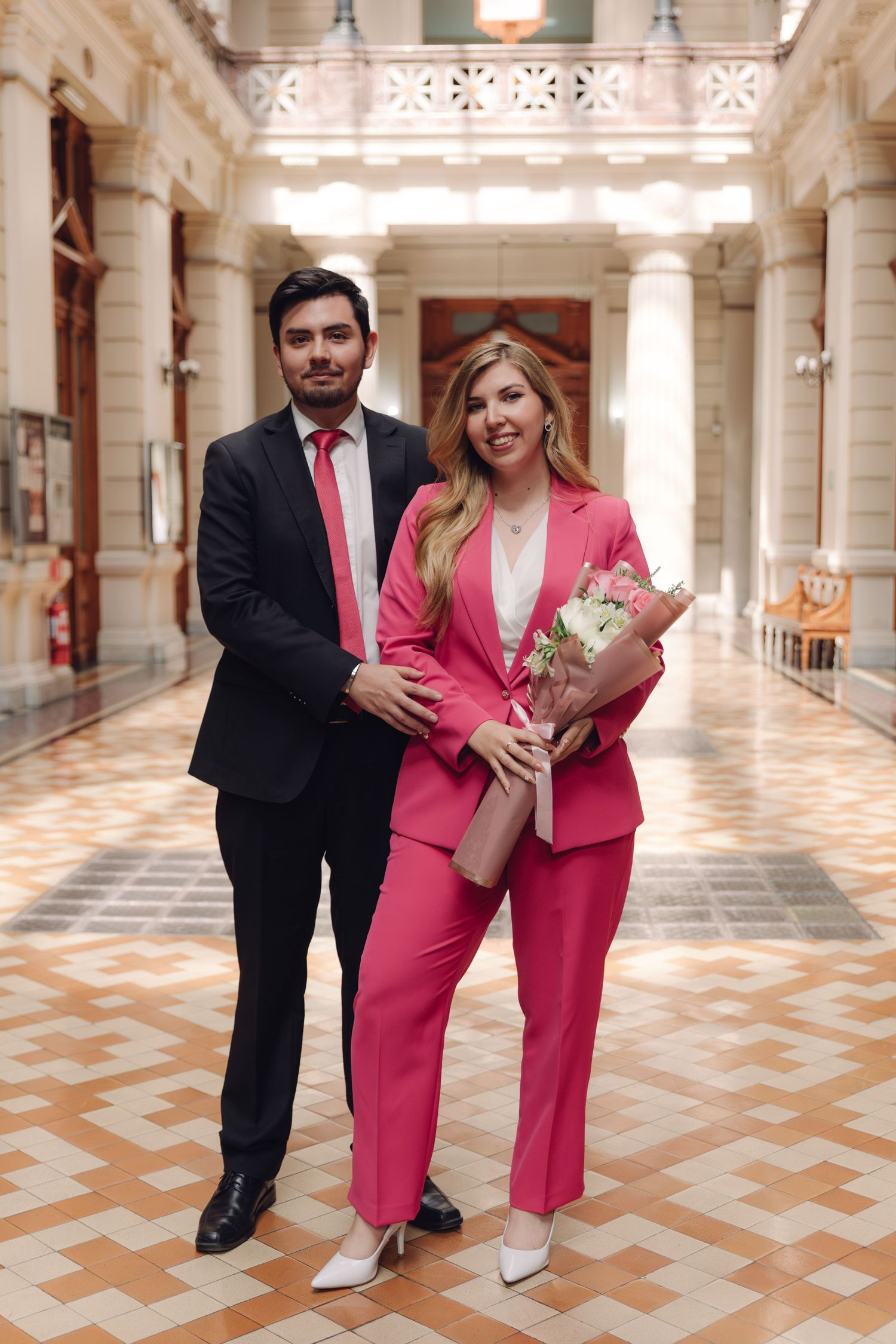 Couple in formal attire poses in elegant hall, woman in pink suit holds bouquet of flowers.