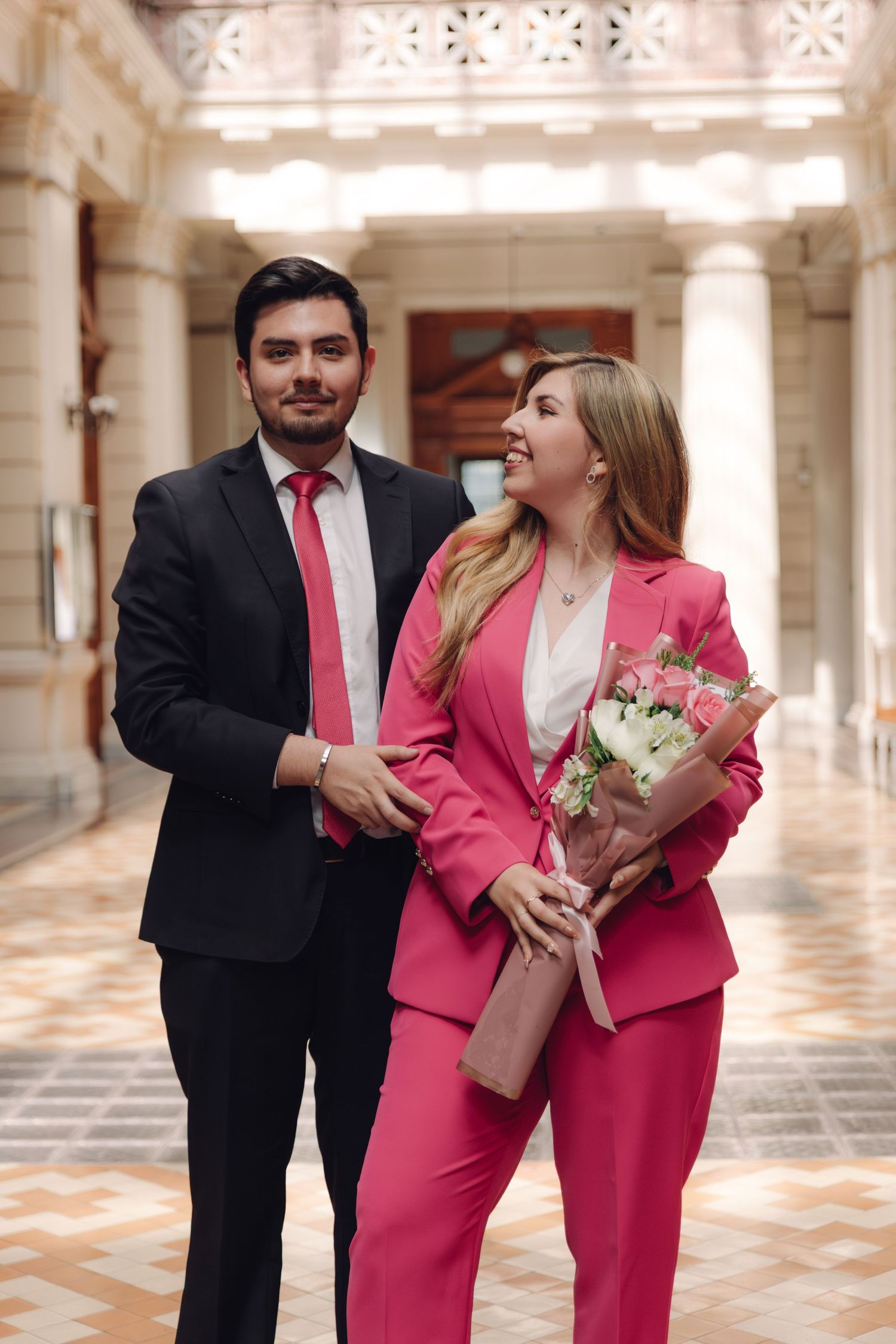 Couple in formal wear walks through ornate columned hallway, woman in pink suit holds bouquet.