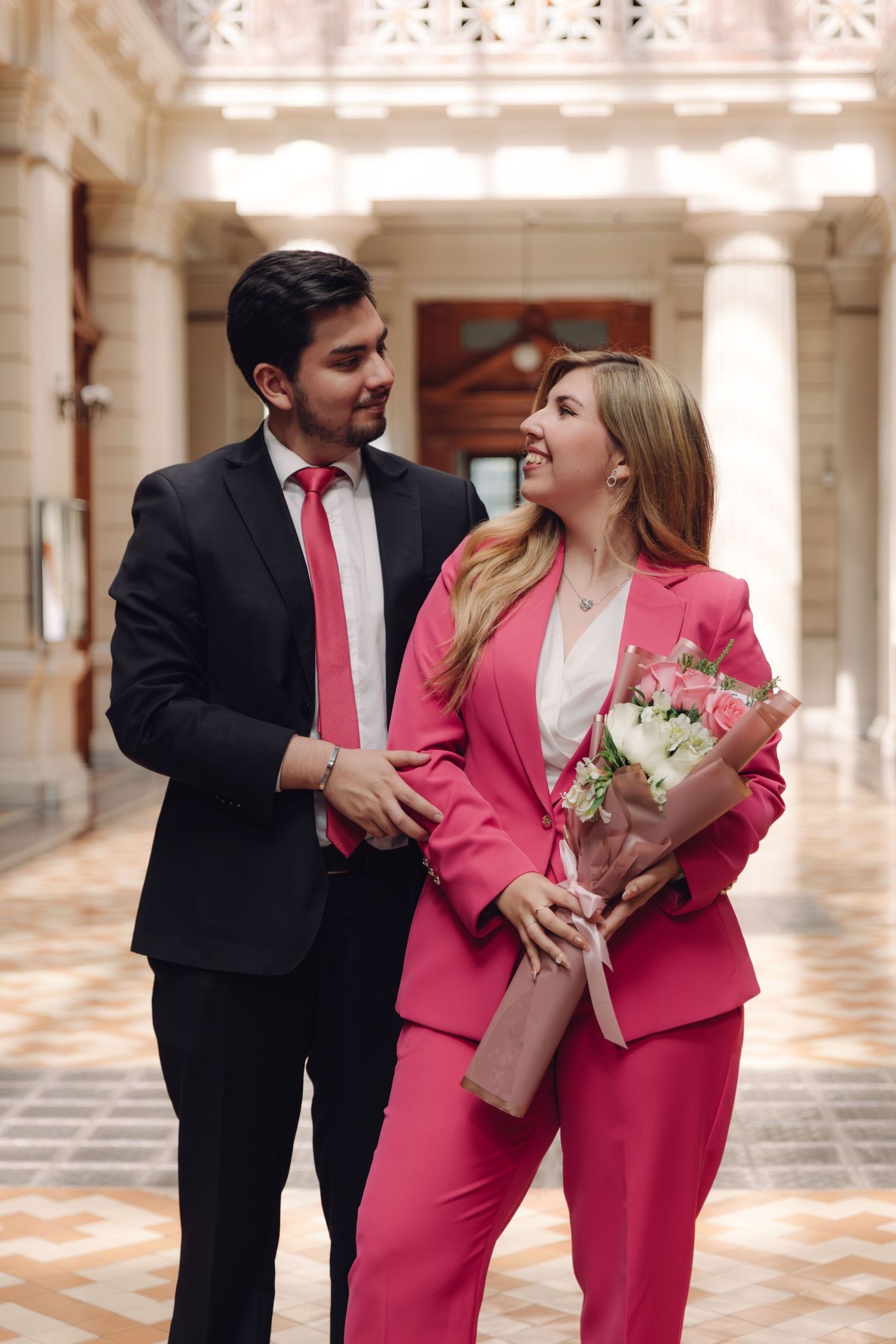 Smiling couple in formal attire share a joyful moment in a grand columned hallway with flowers.