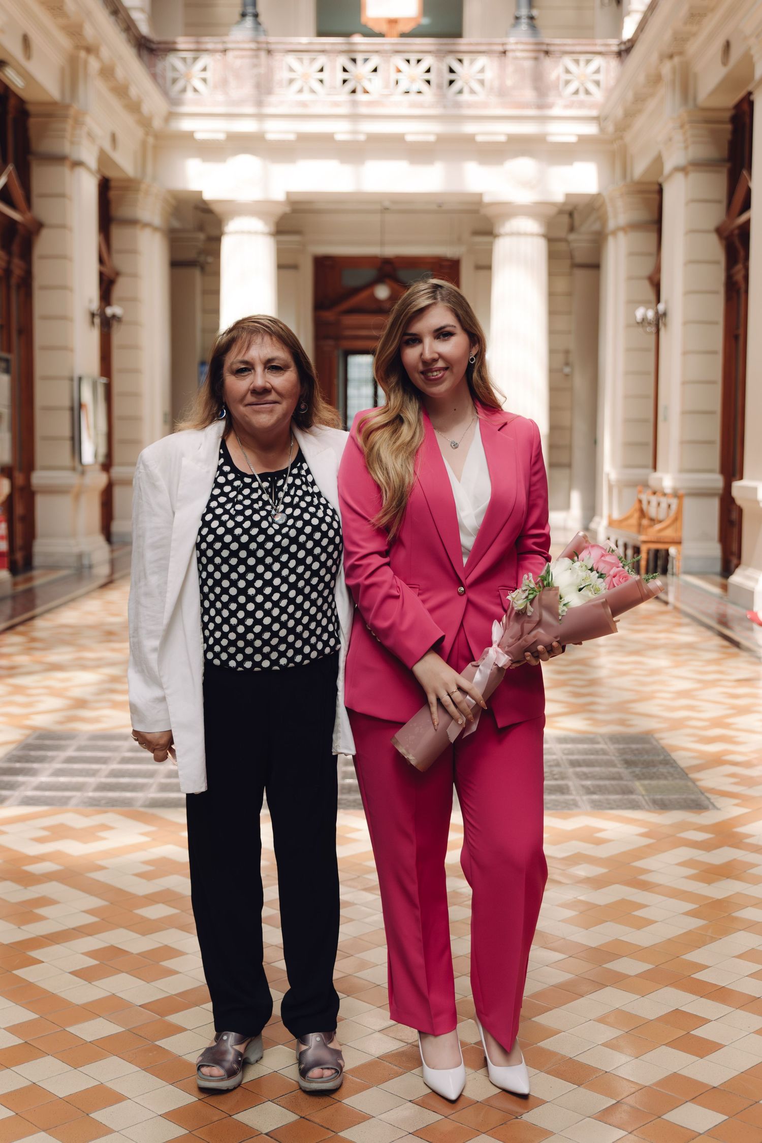 Two women pose together in elegant corridor, younger woman in pink suit holds floral bouquet.